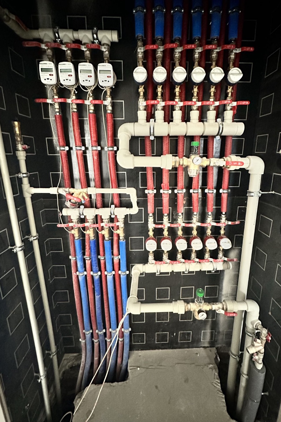 Rozdzielacz ogrzewania podłogowego z licznikami ciepła i manometrami, rury w kolorze czerwonym i niebieskim, montowane na ścianie z czarno-białą wzorzystą tapetą.