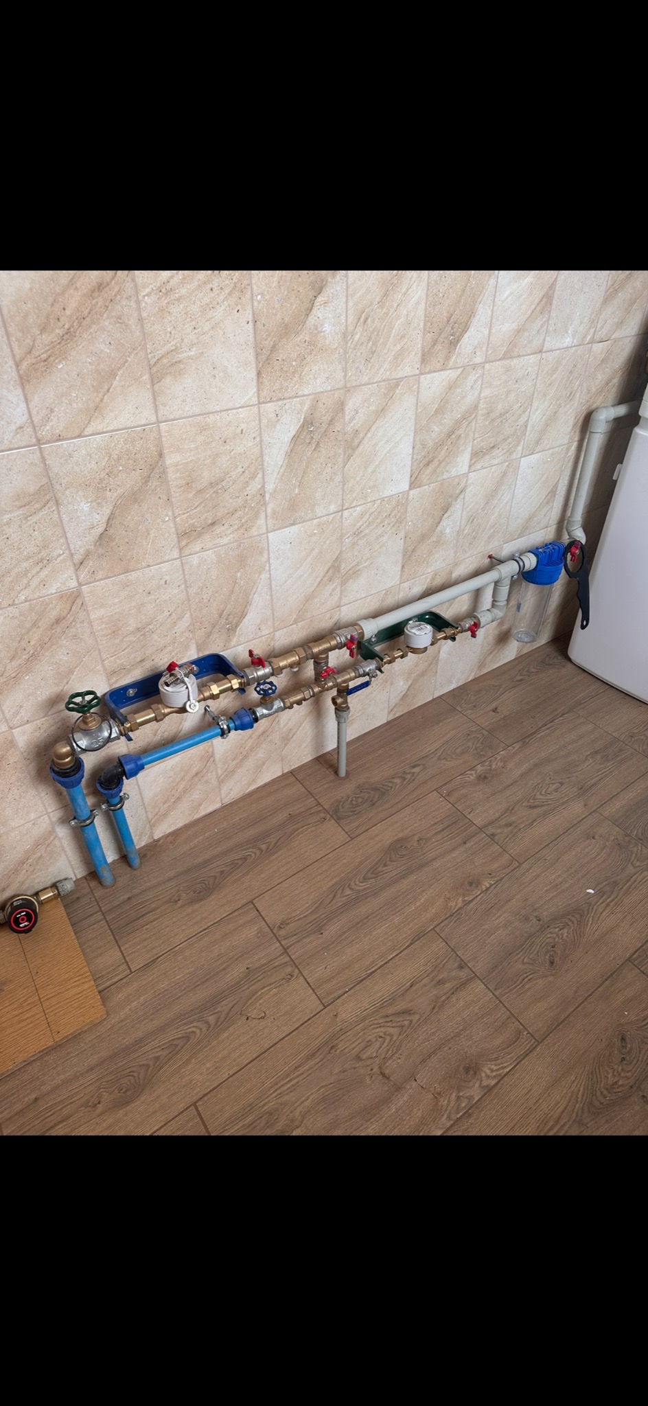 Kompleksowa instalacja wodno-kanalizacyjna z licznymi zaworami, filtrem i manometrem zamontowana przy ścianie z beżowych płytek i drewnopodobnej podłodze.