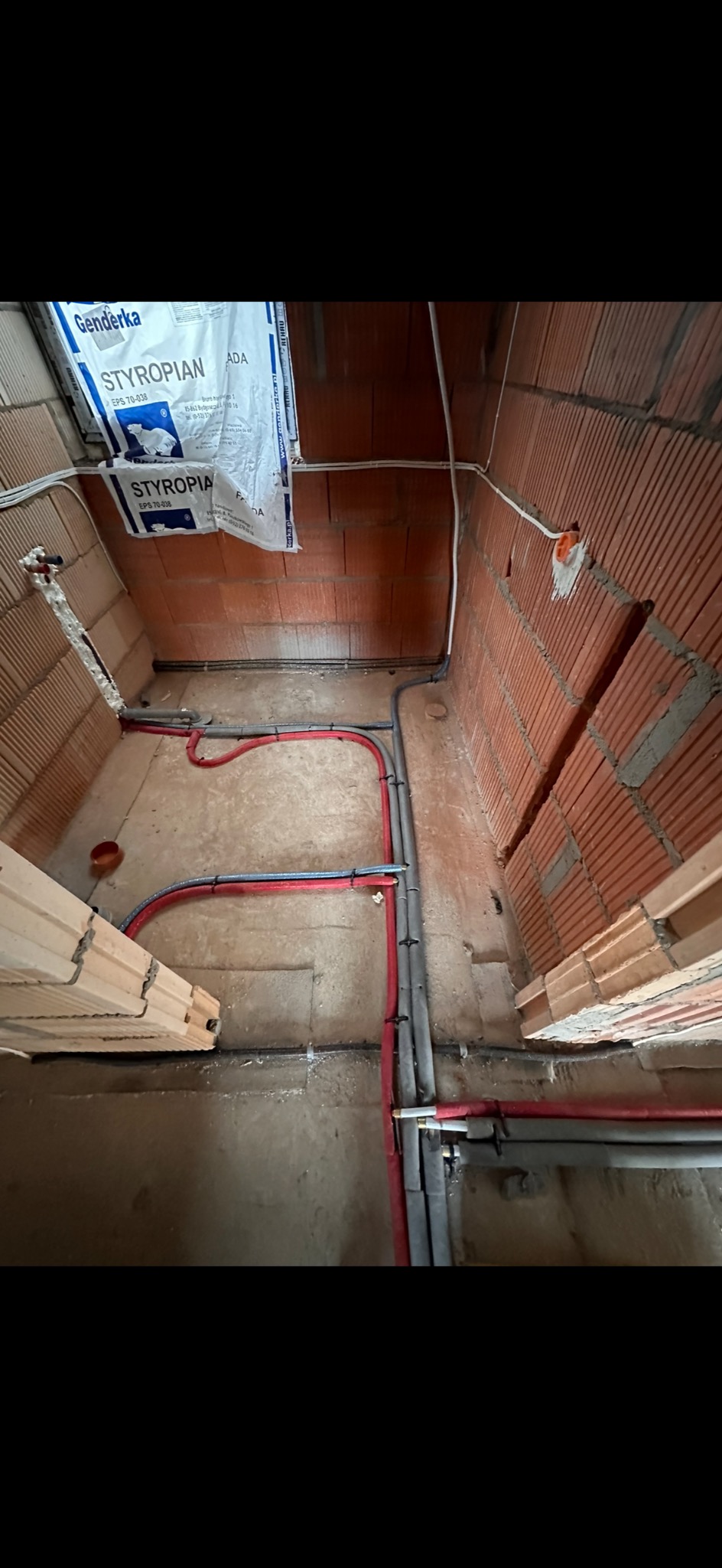 Surowy stan instalacji w pomieszczeniu z ceglanymi ścianami: rury grzewcze w izolacji czerwonej i niebieskiej, szare rury kanalizacyjne, widoczne puszki elektryczne i opakowanie styropianu.