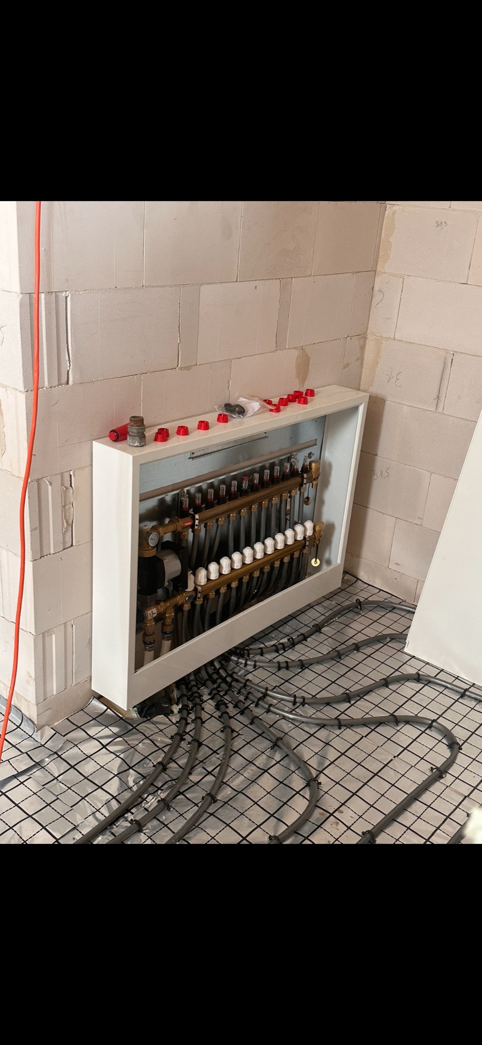 Rozdzielacz ogrzewania podłogowego z widocznymi rurami rozprowadzonymi na folii izolacyjnej, w tle ściana z betonu komórkowego.