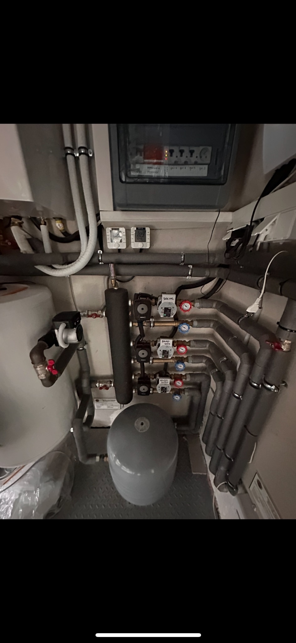 Złożony system rur grzewczych i zaworów w piwnicy, widoczne izolowane rury, zbiornik wyrównawczy, pompy obiegowe i panel sterowania, szare tło.