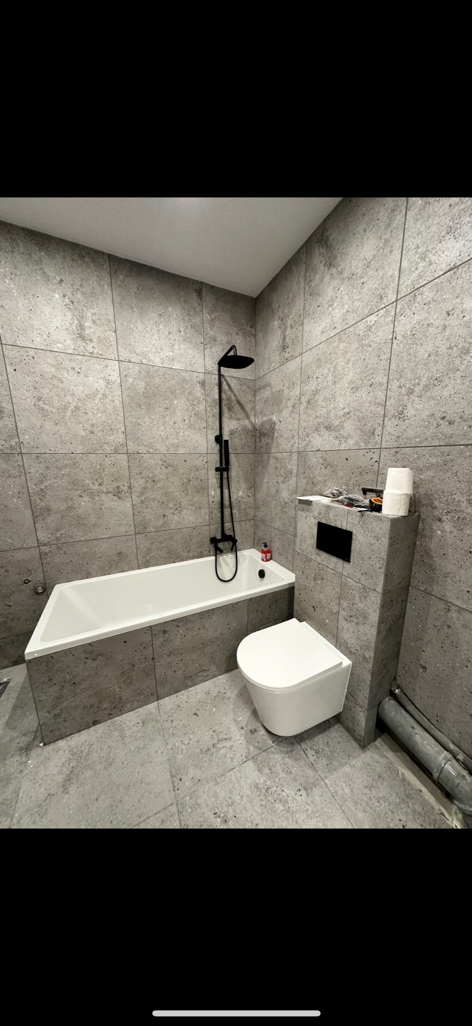 Wykończona łazienka w Środzie Wielkopolskiej z wanną, toaletą podwieszaną i deszczownicą, wyłożona szarymi płytkami imitującymi beton, z widocznymi rurami kanalizacyjnymi.