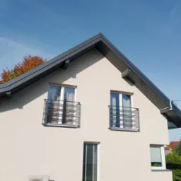 Dwukondygnacyjny dom z jasną elewacją i szarym dachem dwuspadowym, widoczne okna z balkonami francuskimi, jesienne drzewa w tle, błękitne niebo.