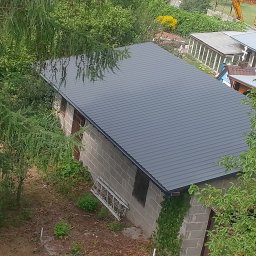 Js-konstrukcje - Szary dach z blachy trapezowej na murowanym budynku gospodarczym, widziany z góry, z fragmentami drzew i drabiny opartą o ścianę. W tle widoczny wykop i koparka.