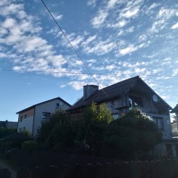 Js-konstrukcje - Dom w trakcie remontu dachu, widoczne rusztowania i częściowo zdjęte pokrycie. Błękitne niebo z chmurami w tle. Ujęcie zza drewnianego płotu.