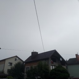 Js-konstrukcje - Dom jednorodzinny z nowym, ciemnobrązowym dachem z blachodachówki, widoczny na tle pochmurnego nieba. Widoczne przewody elektryczne nad budynkami.