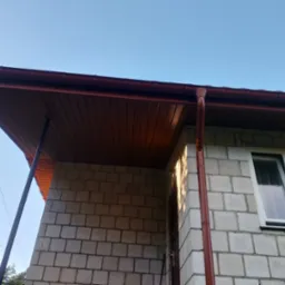 Nowa, brązowa rynna i podbitka dachowa na tle błękitnego nieba w Czarnej Białostockiej. Widok z dołu na fragment budynku z oknem.