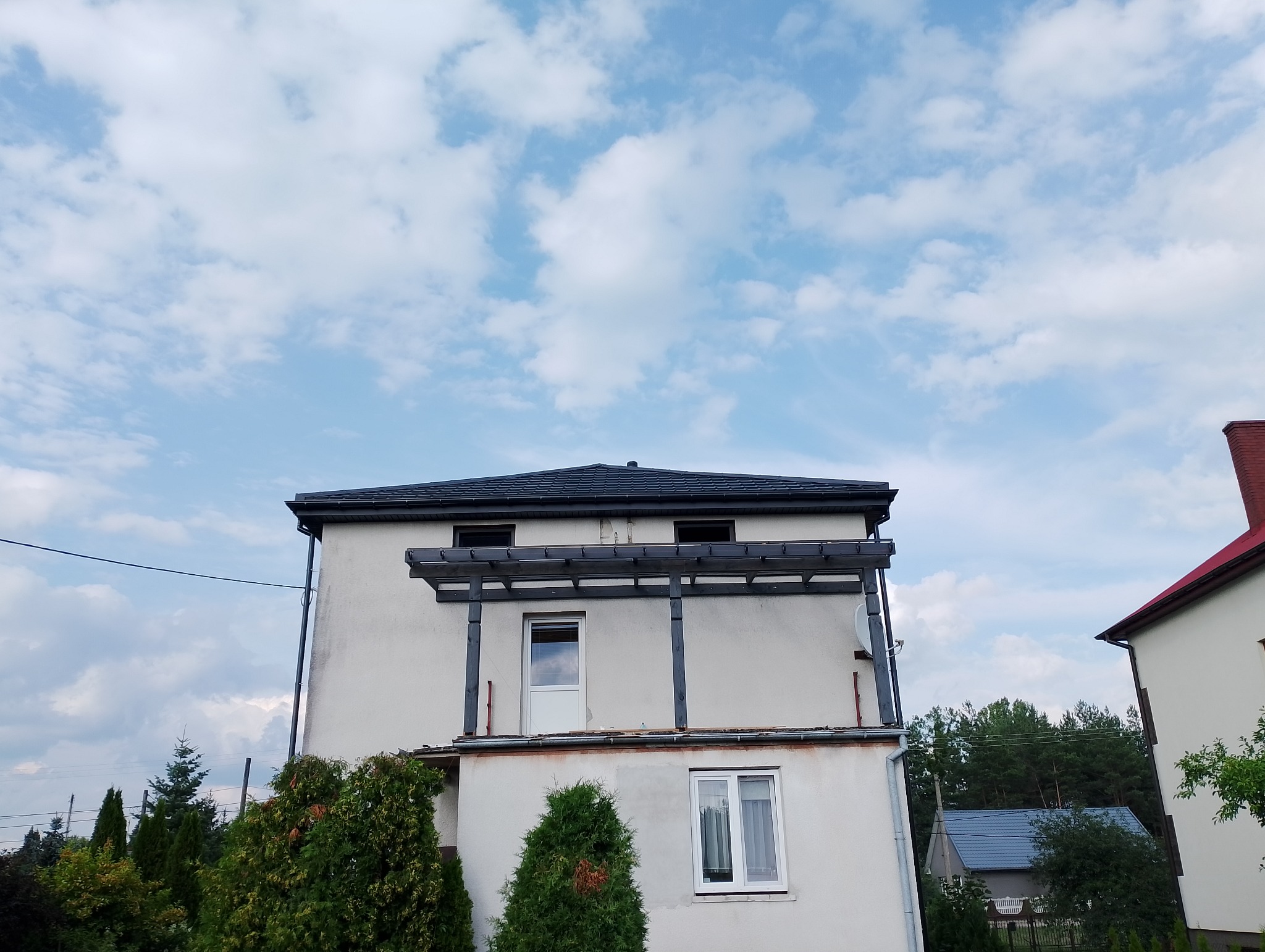 Dwupiętrowy dom z ciemnoszarym dachem z dachówki ceramicznej, widoczna drewniana konstrukcja balkonu i biała elewacja, na tle błękitnego nieba z obłokami.