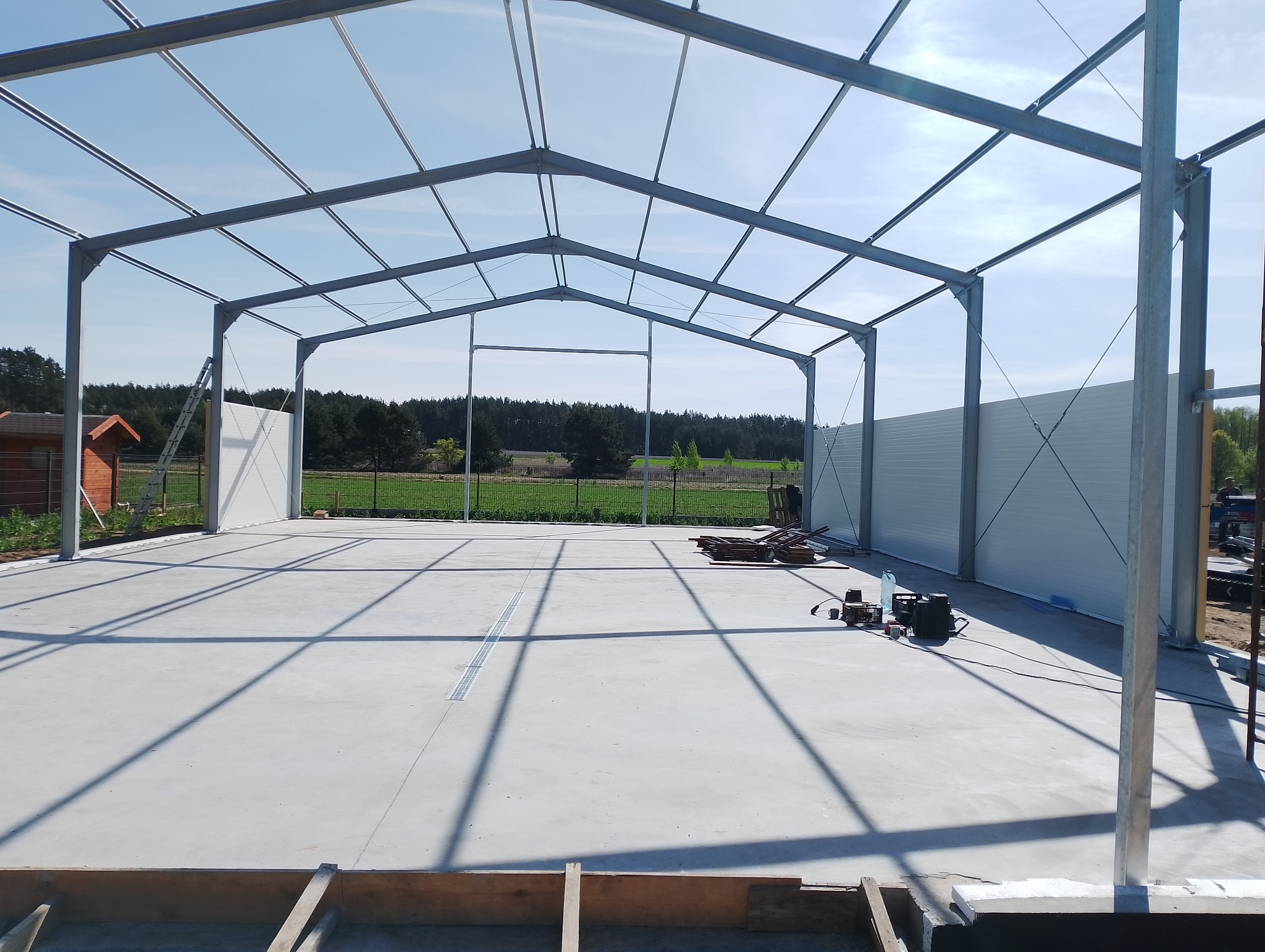 Konstrukcja stalowa hali w trakcie budowy, widoczne elementy szkieletu i fragmenty ścian z płyt warstwowych. Na pierwszym planie elementy drewniane, w tle zieleń.