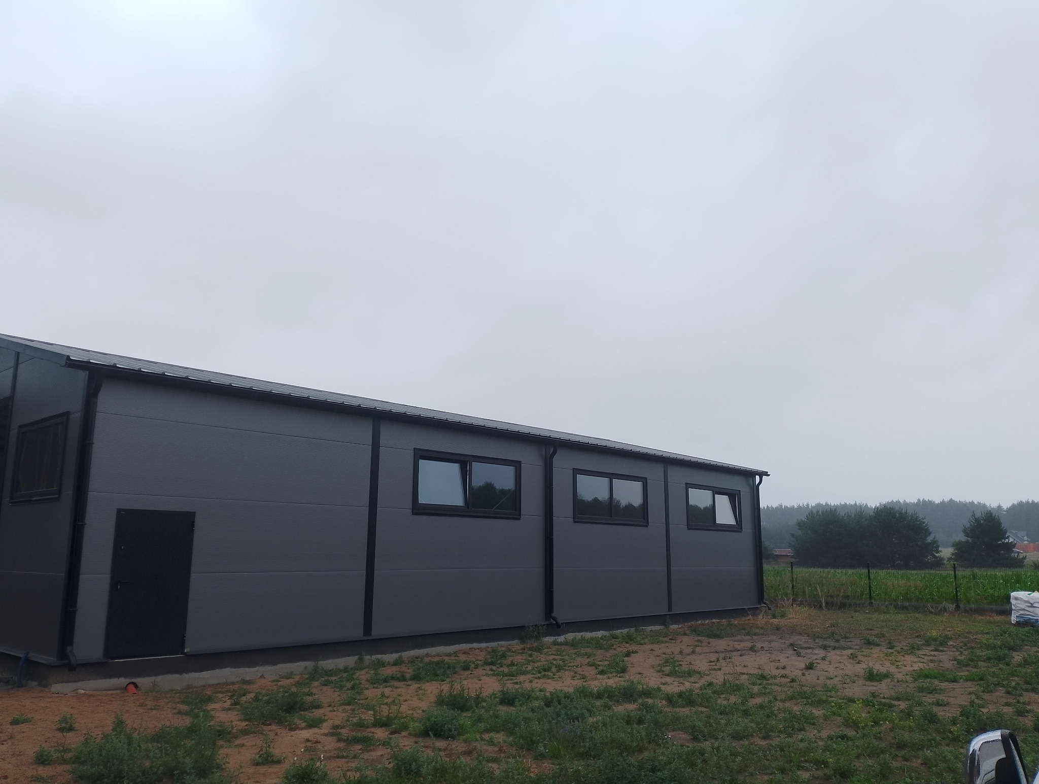 Szara hala z płyt warstwowych, widok z zewnątrz w pochmurny dzień. Czarna stolarka okienna i drzwiowa, obróbki blacharskie w kolorze dachu.
