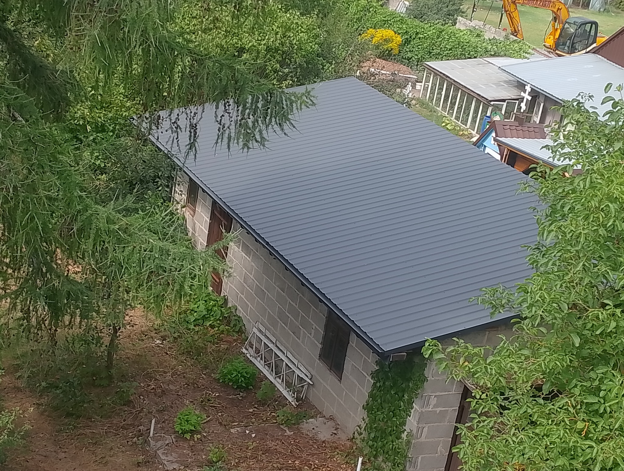 Szary dach z blachy trapezowej na murowanym budynku gospodarczym, widziany z góry, z fragmentami drzew i drabiny opartą o ścianę. W tle widoczny wykop i koparka.