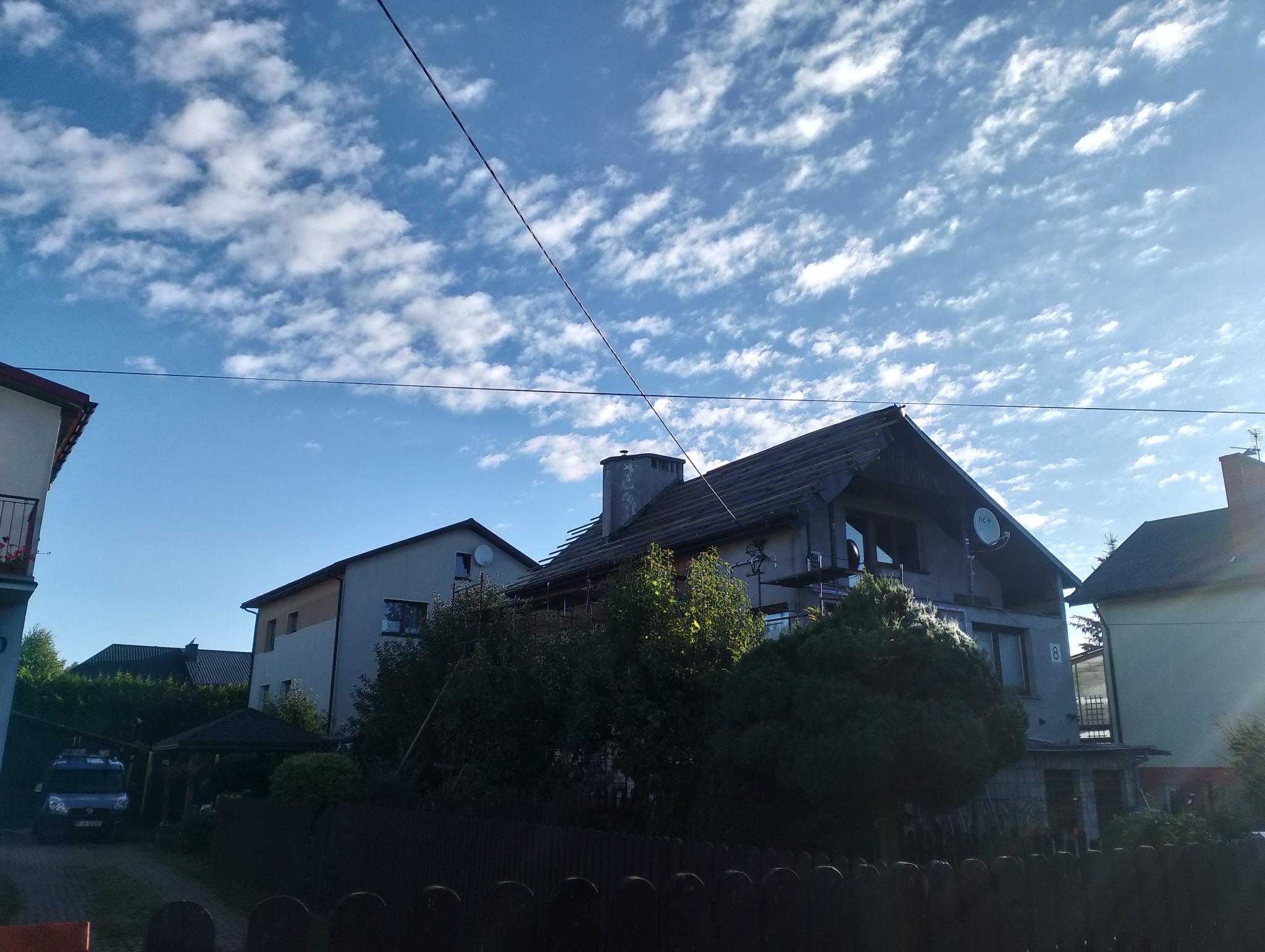 Dom w trakcie remontu dachu, widoczne rusztowania i częściowo zdjęte pokrycie. Błękitne niebo z chmurami w tle. Ujęcie zza drewnianego płotu.