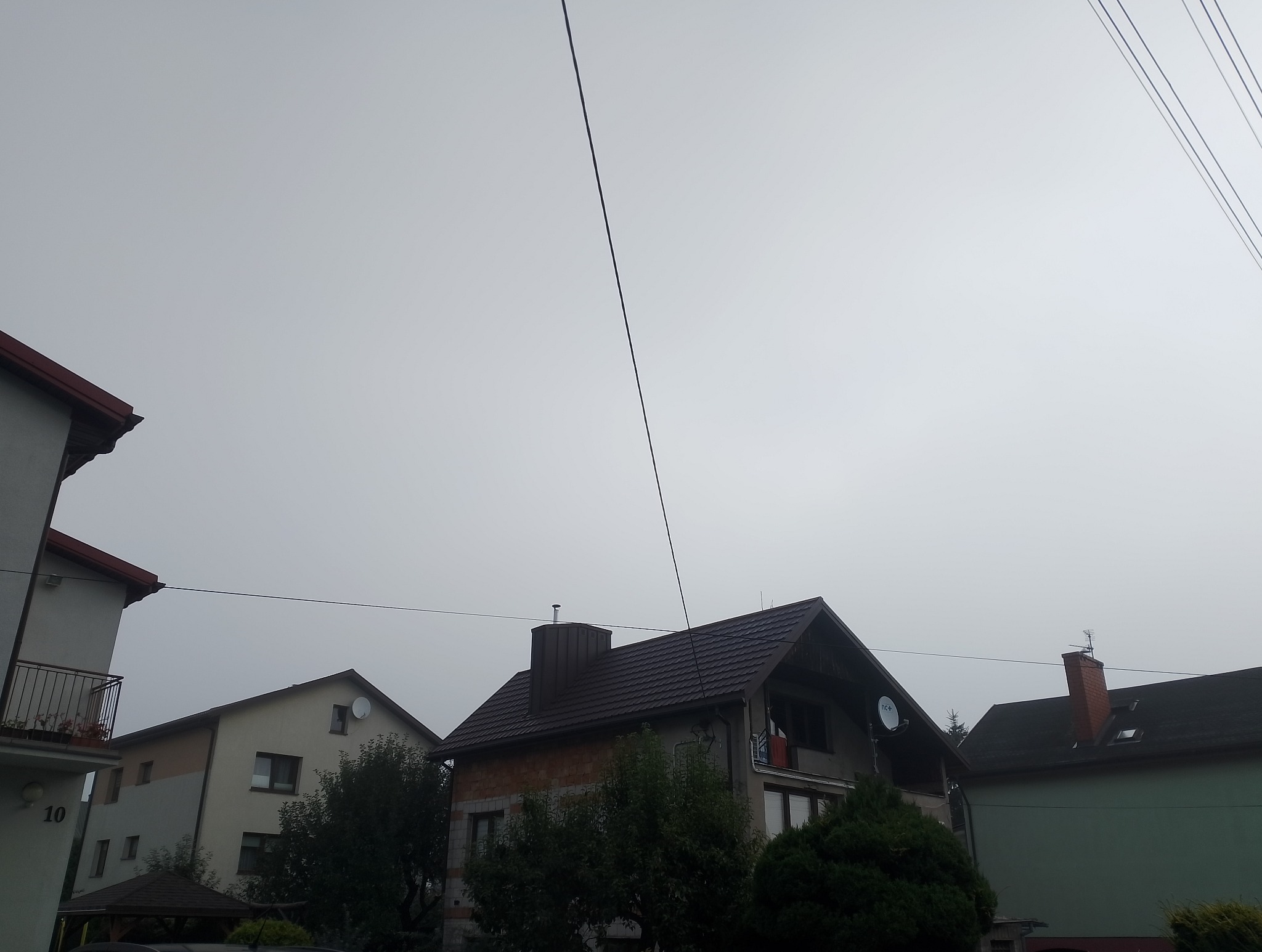 Dom jednorodzinny z nowym, ciemnobrązowym dachem z blachodachówki, widoczny na tle pochmurnego nieba. Widoczne przewody elektryczne nad budynkami.