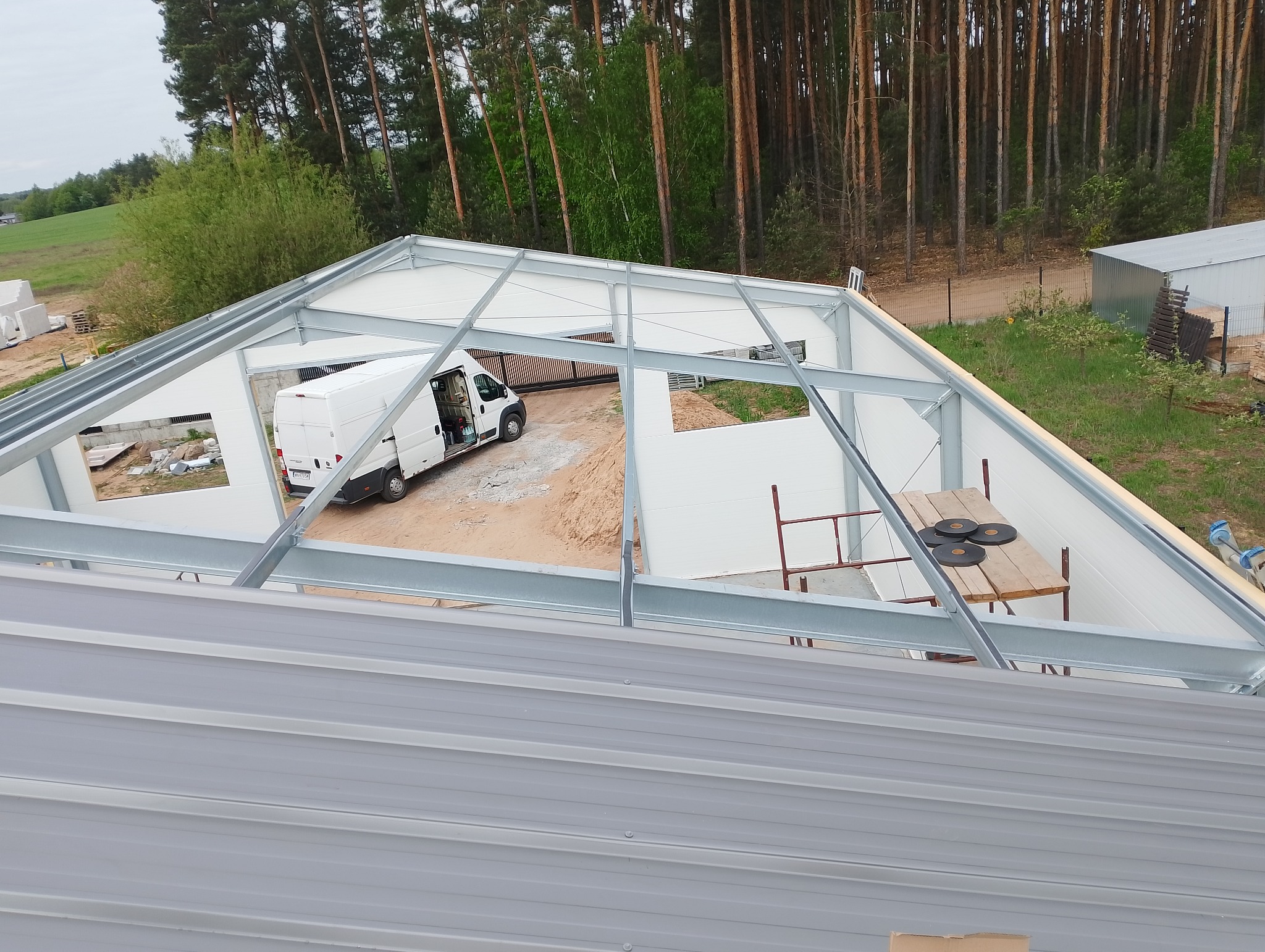 Montaż konstrukcji stalowej hali z płyt warstwowych, widok z góry na częściowo pokryty dach. Biały bus w tle. Etap budowy.