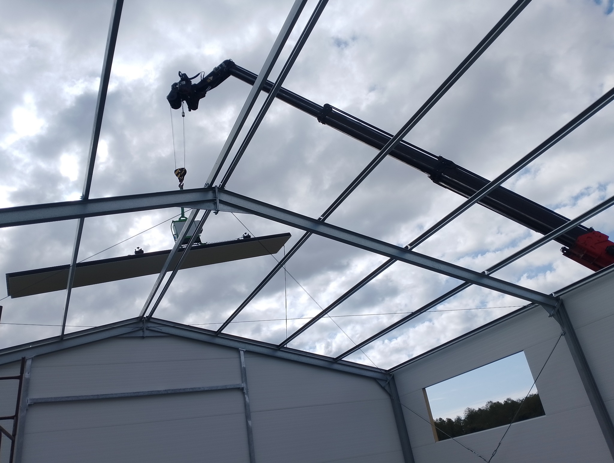Montaż płyty warstwowej w hali stalowej za pomocą dźwigu. Widok od wewnątrz, na tle pochmurnego nieba. Konstrukcja z metalowych belek.