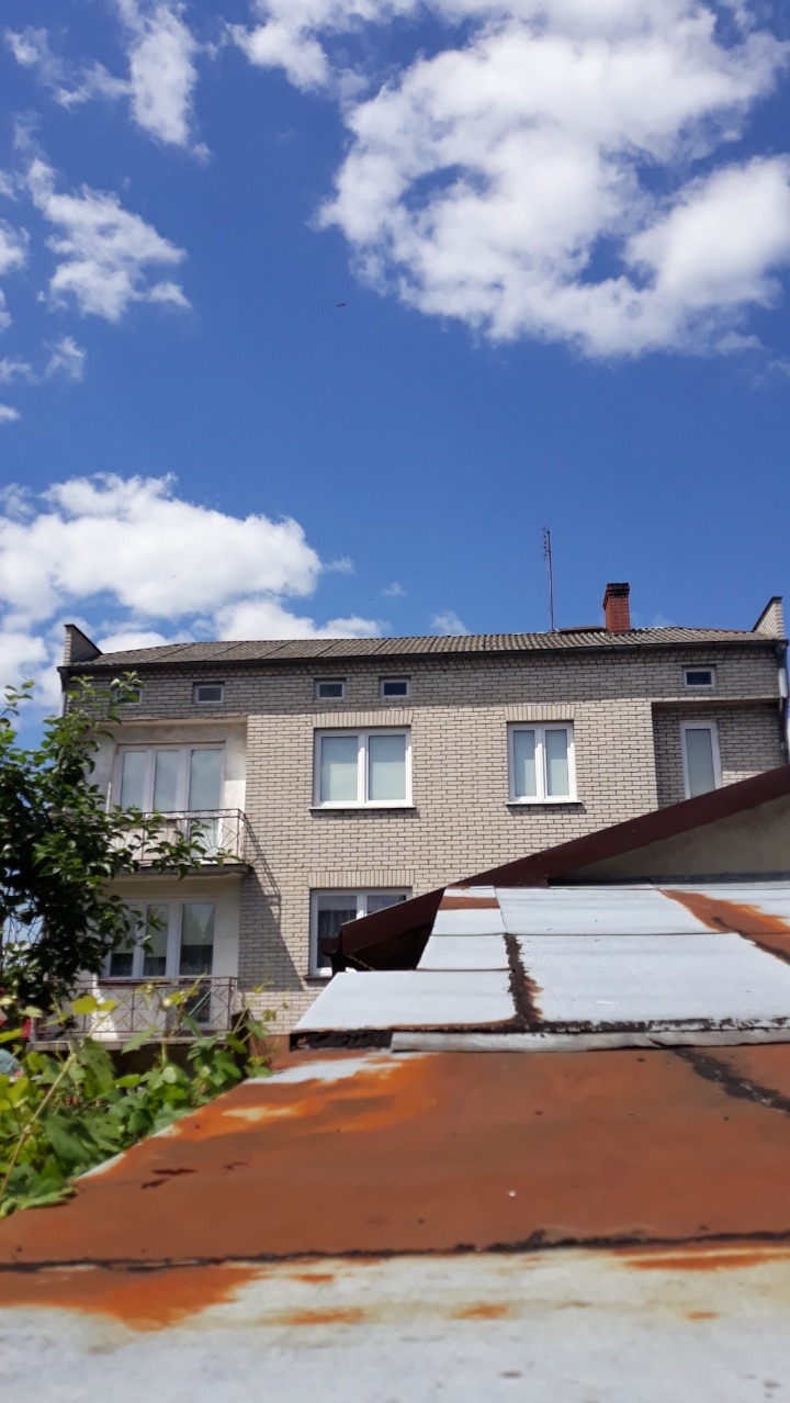 Dwupiętrowy budynek z cegły pod błękitnym niebem z chmurami, widziany z perspektywy dachu pokrytego rdzawą blachą. Widoczny komin i antena.