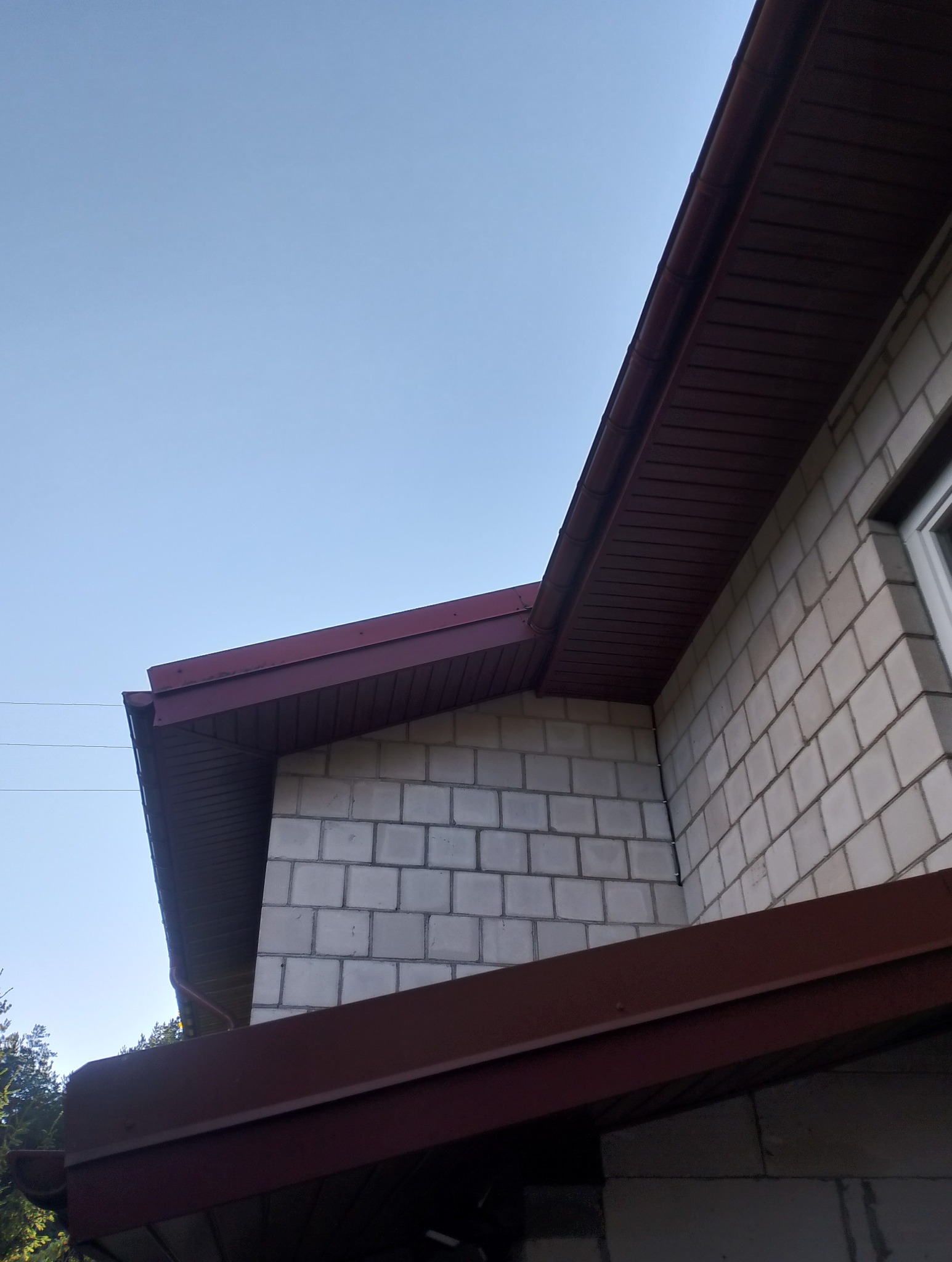 Fragment budynku z dachem pokrytym blachą w kolorze ciemnej czerwieni, widoczne orynnowanie i ściana z białych bloczków. Ujęcie z dołu, niebieskie niebo w tle.