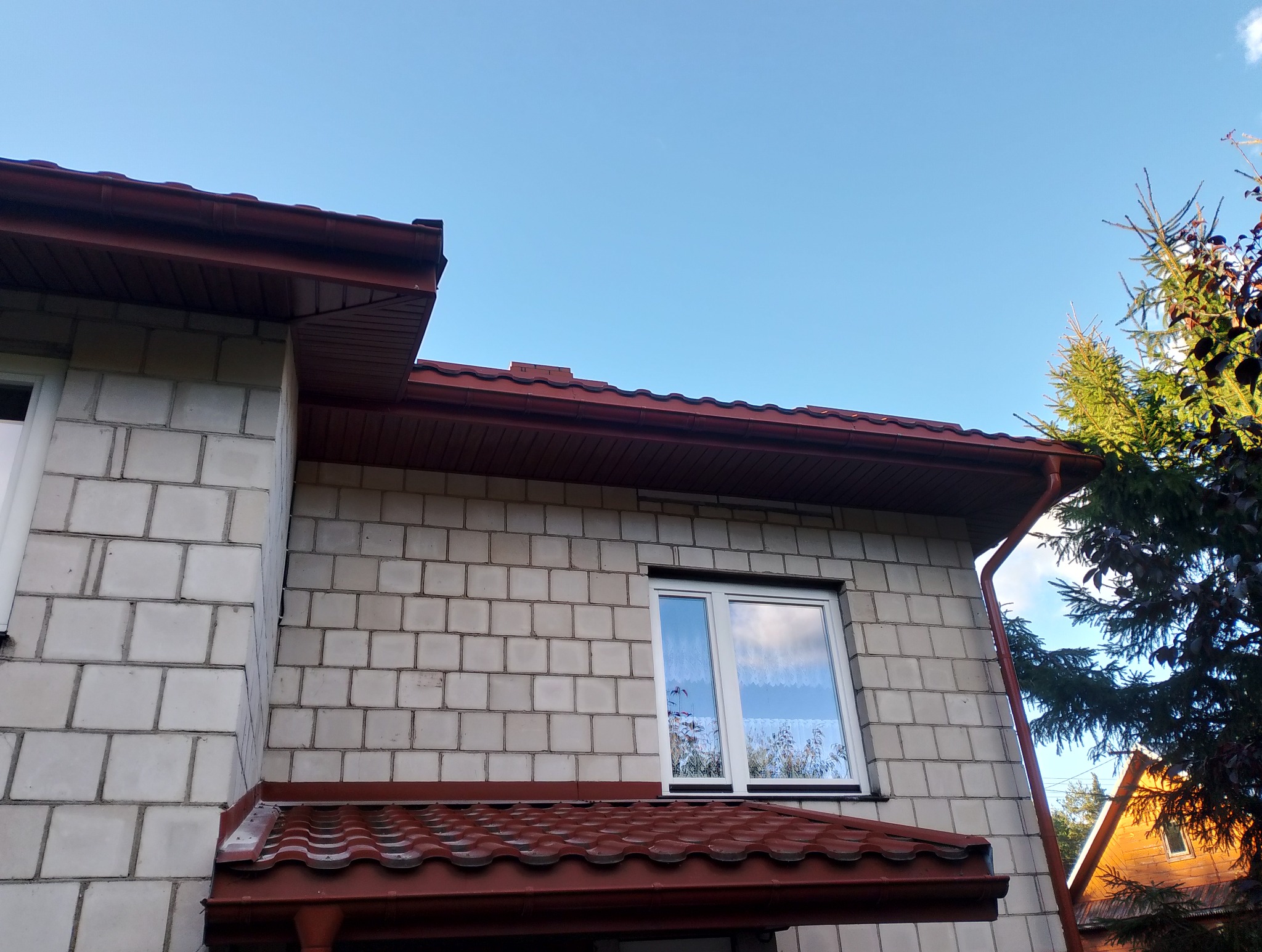 Dom z jasnej cegły z nową, czerwoną dachówką i rynnami. Widoczne okno i fragment drzewa. Ujęcie z dołu, niebo w tle.