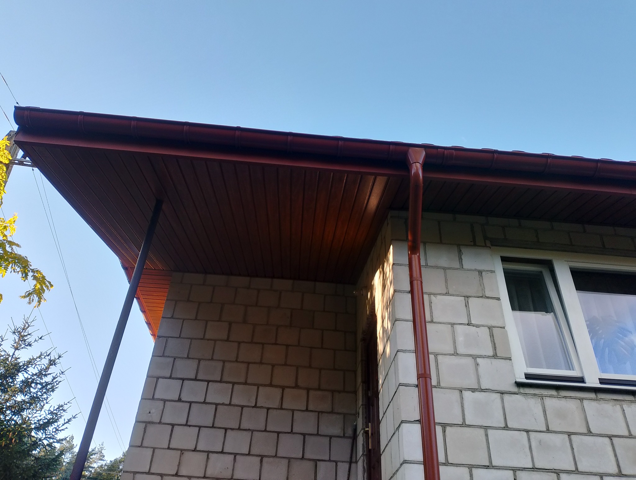 Nowa, brązowa rynna i podbitka dachowa na tle błękitnego nieba w Czarnej Białostockiej. Widok z dołu na fragment budynku z oknem.