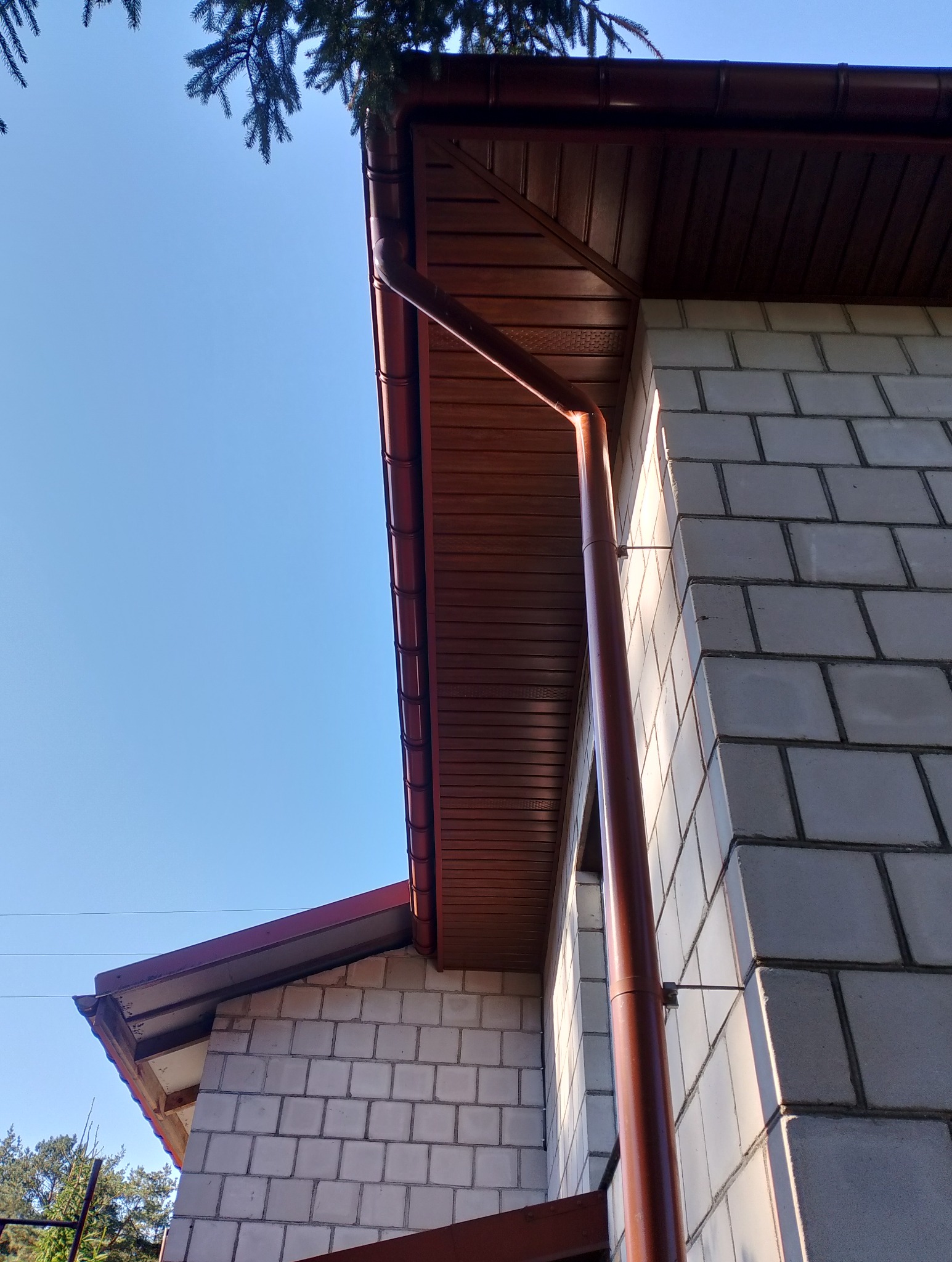 Fragment budynku z brązowymi rynnami i podbitką dachową, widoczne bloczki betonowe i fragment iglastego drzewa na tle błękitnego nieba.