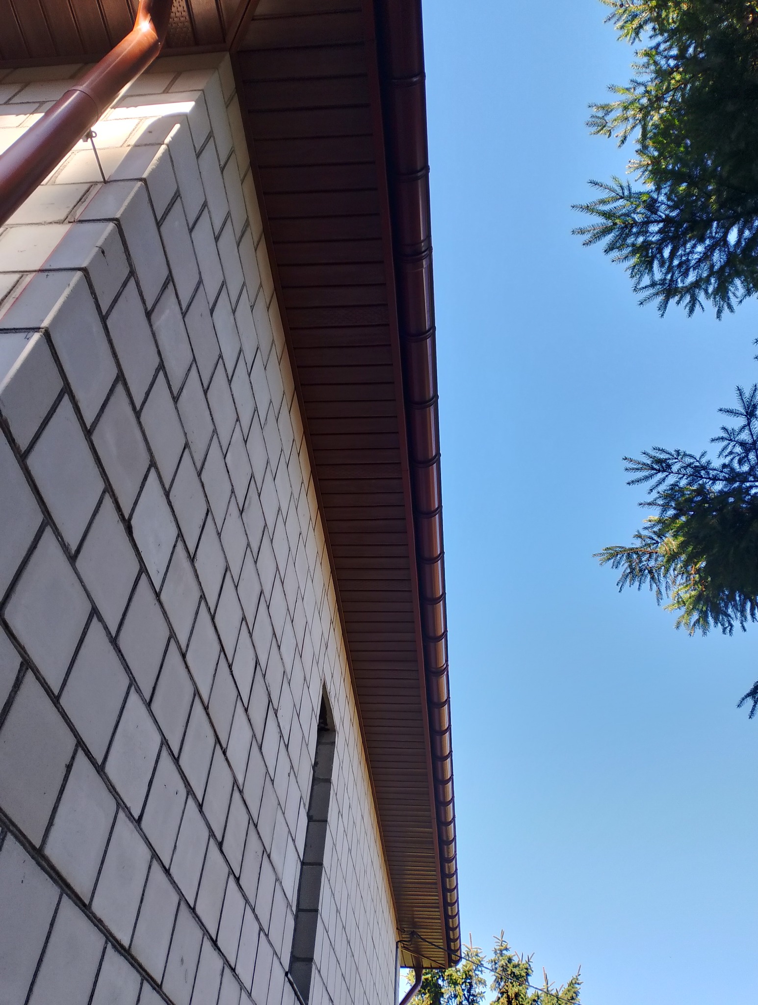 Ściana budynku z białej cegły, brązowe orynnowanie i podbitka dachowa na tle błękitnego nieba i zieleni drzew. Ujęcie z dołu.