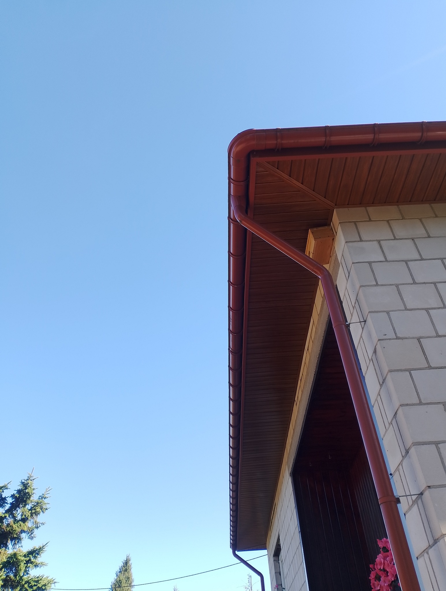 Fragment budynku z brązowym dachem i rynnami na tle błękitnego nieba. Widoczne białe bloczki i zielone drzewa. Ujęcie z dołu, podkreślające detale wykończenia dachu.