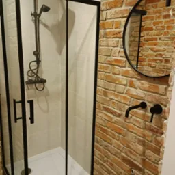 Kabina prysznicowa z czarnymi ramami i armaturą na tle ściany z cegły, okrągłe lustro w czarnej ramie, drewnopodobna podłoga.