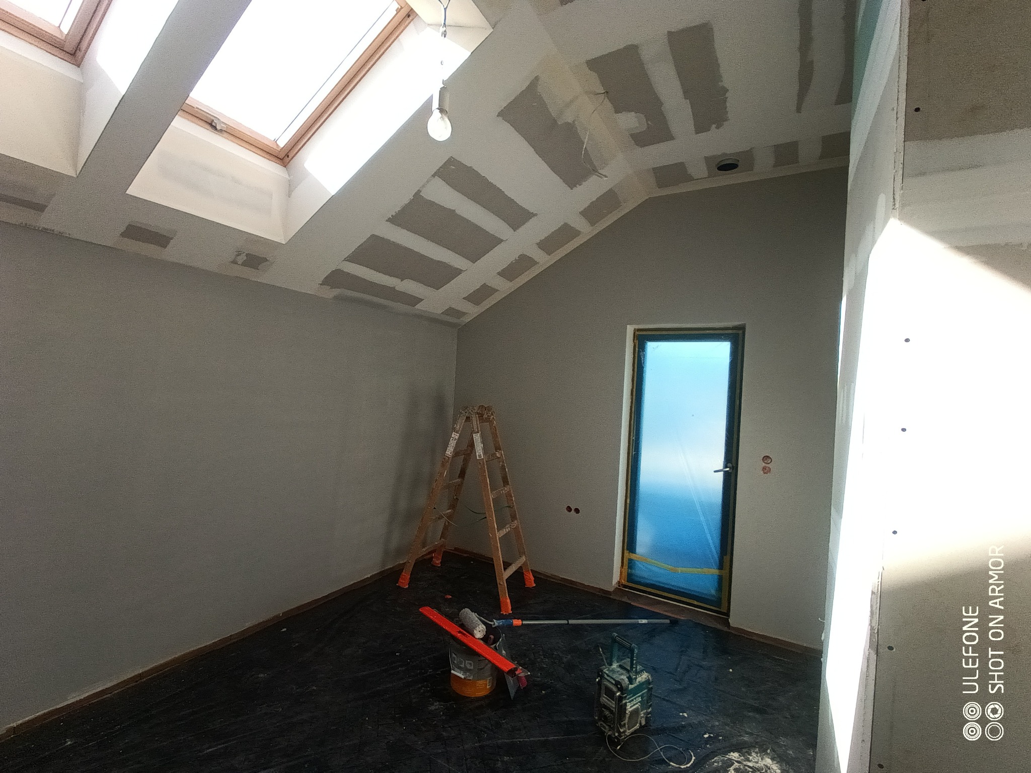 Poddasze w trakcie prac wykończeniowych: widoczne okna dachowe, szpachlowane łączenia płyt na suficie, drabina malarska, wiadro z narzędziami i zabezpieczone folią drzwi.