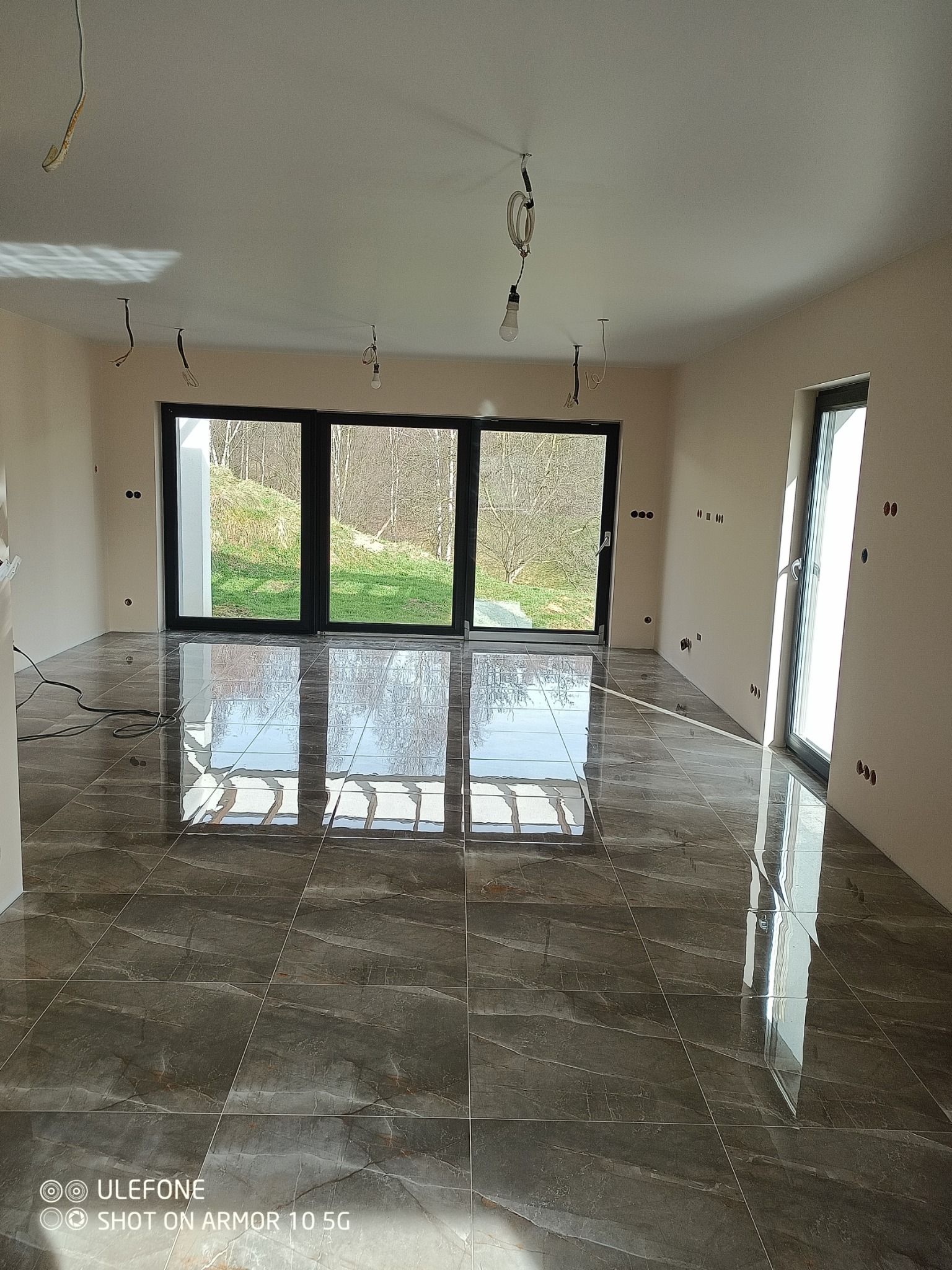 Wnętrze pokoju z ciemnymi, błyszczącymi panelami podłogowymi, dużymi oknami z widokiem na zewnątrz, przygotowane do dalszych prac wykończeniowych, widoczne wystające przewody elektryczne z sufitu.