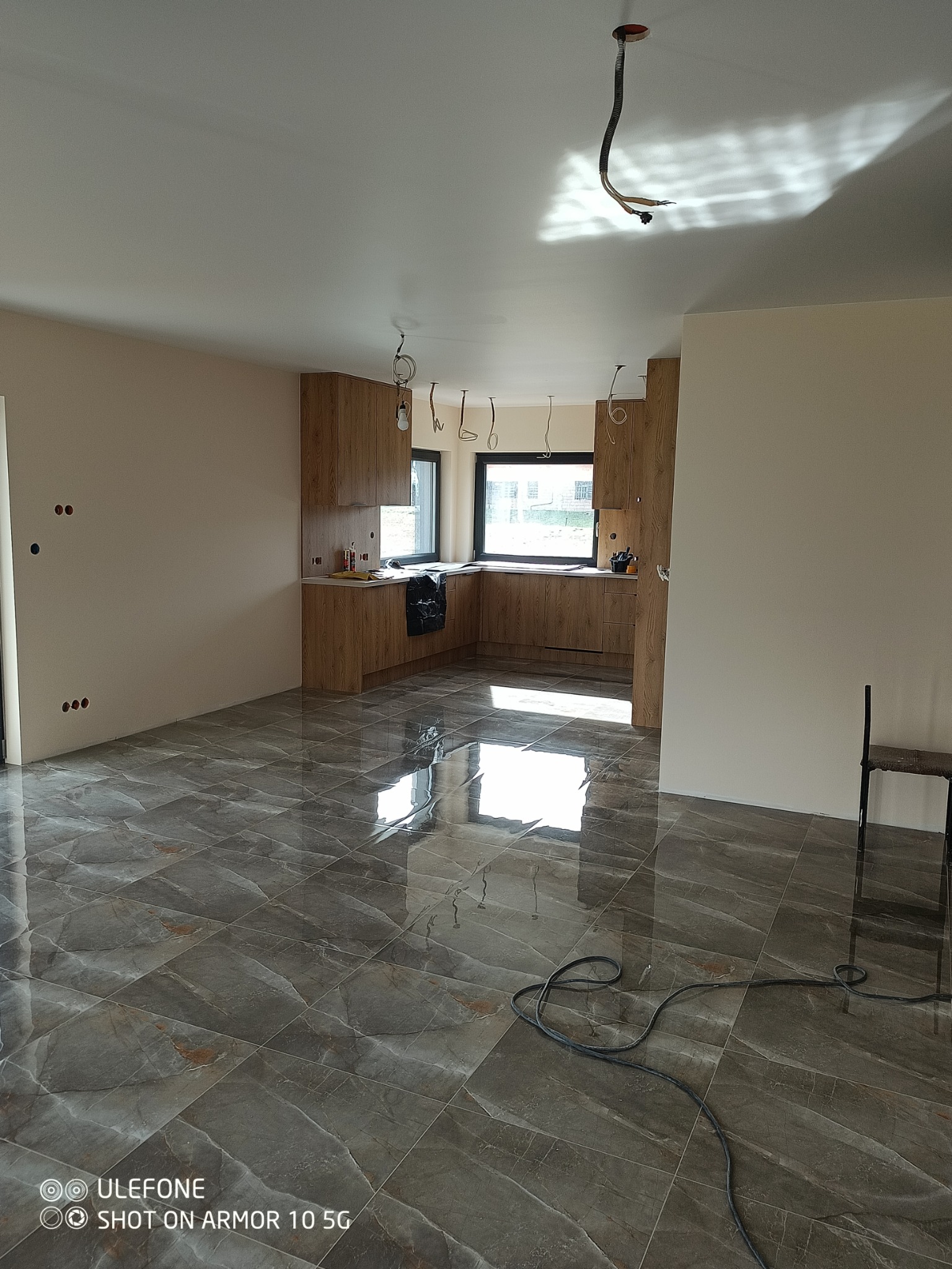 Wnętrze w trakcie wykańczania: położona marmurkowa podłoga, surowe ściany z widocznymi puszkami elektrycznymi, zabudowa kuchenna z drewna, wiszące kable oświetleniowe.