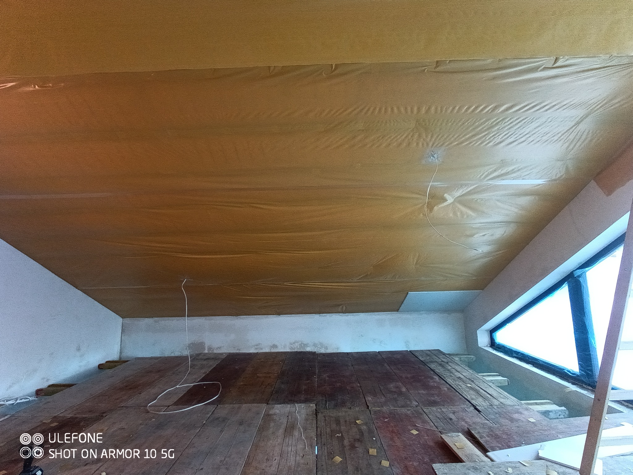 Poddasze w trakcie prac izolacyjnych, widoczna folia ochronna na izolacji, drewniana podłoga i nietypowe trójkątne okno w tle.