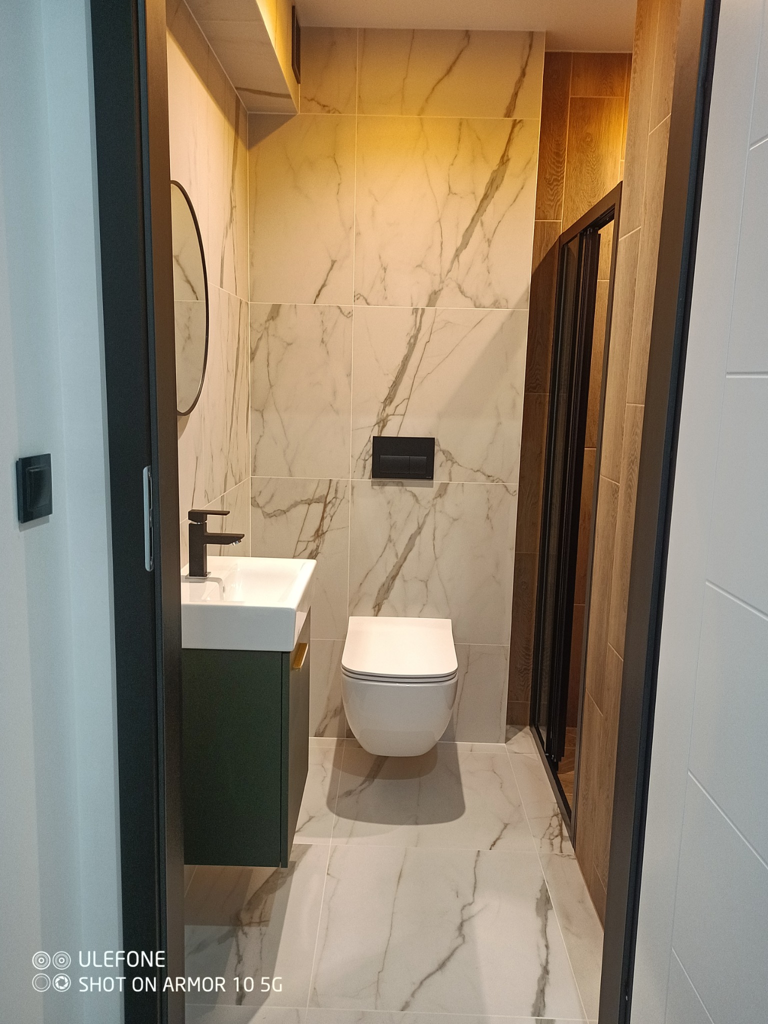 Nowoczesna łazienka z marmurowymi płytkami, wiszącą toaletą, umywalką na szafce w odcieniach zieleni i czarną armaturą, oświetlenie LED w suficie.