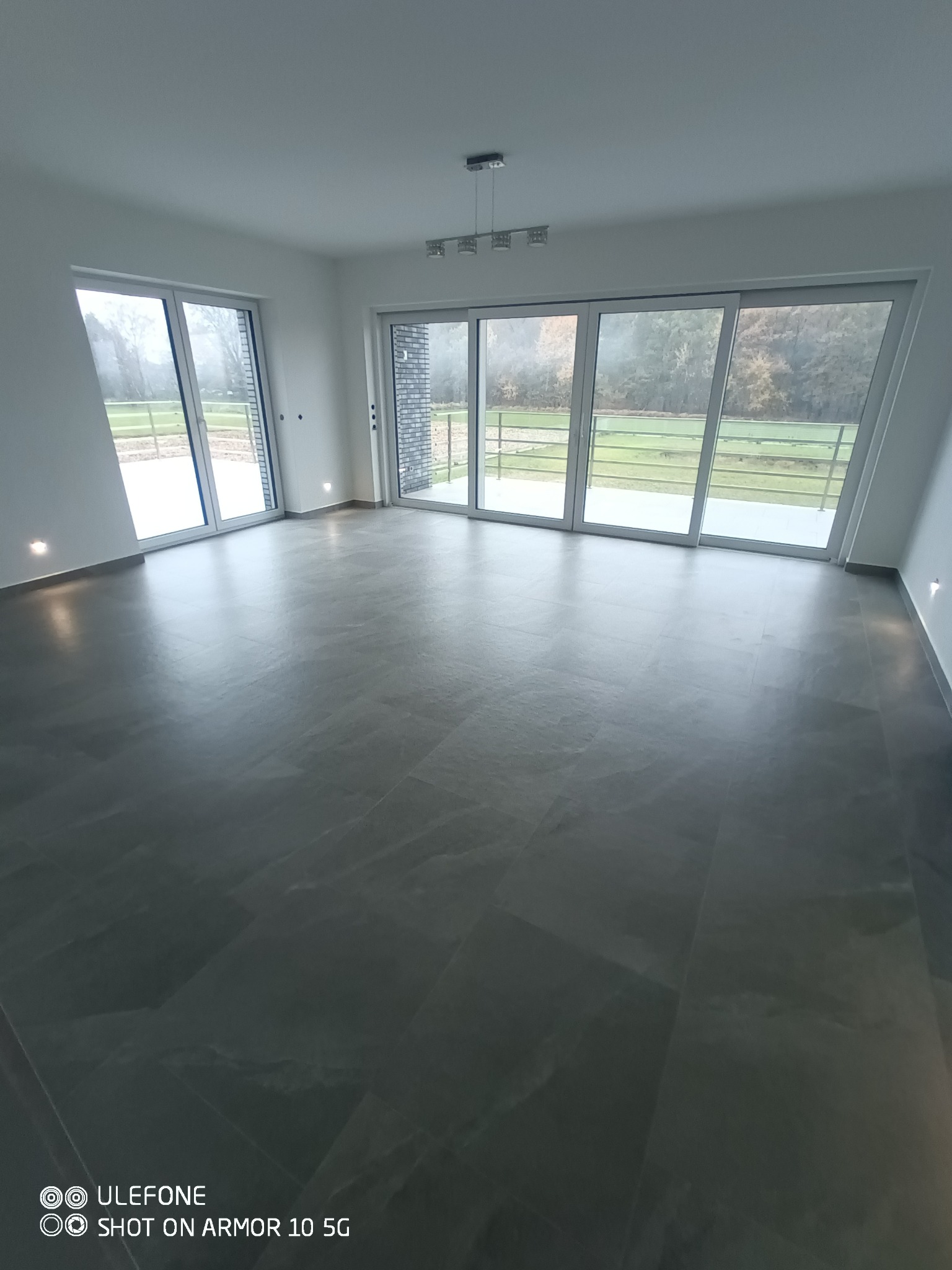 Nowoczesne, przestronne wnętrze z szarą, błyszczącą podłogą z płytek, dużymi oknami przesuwnymi wychodzącymi na zewnątrz i minimalistycznym oświetleniem sufitowym.