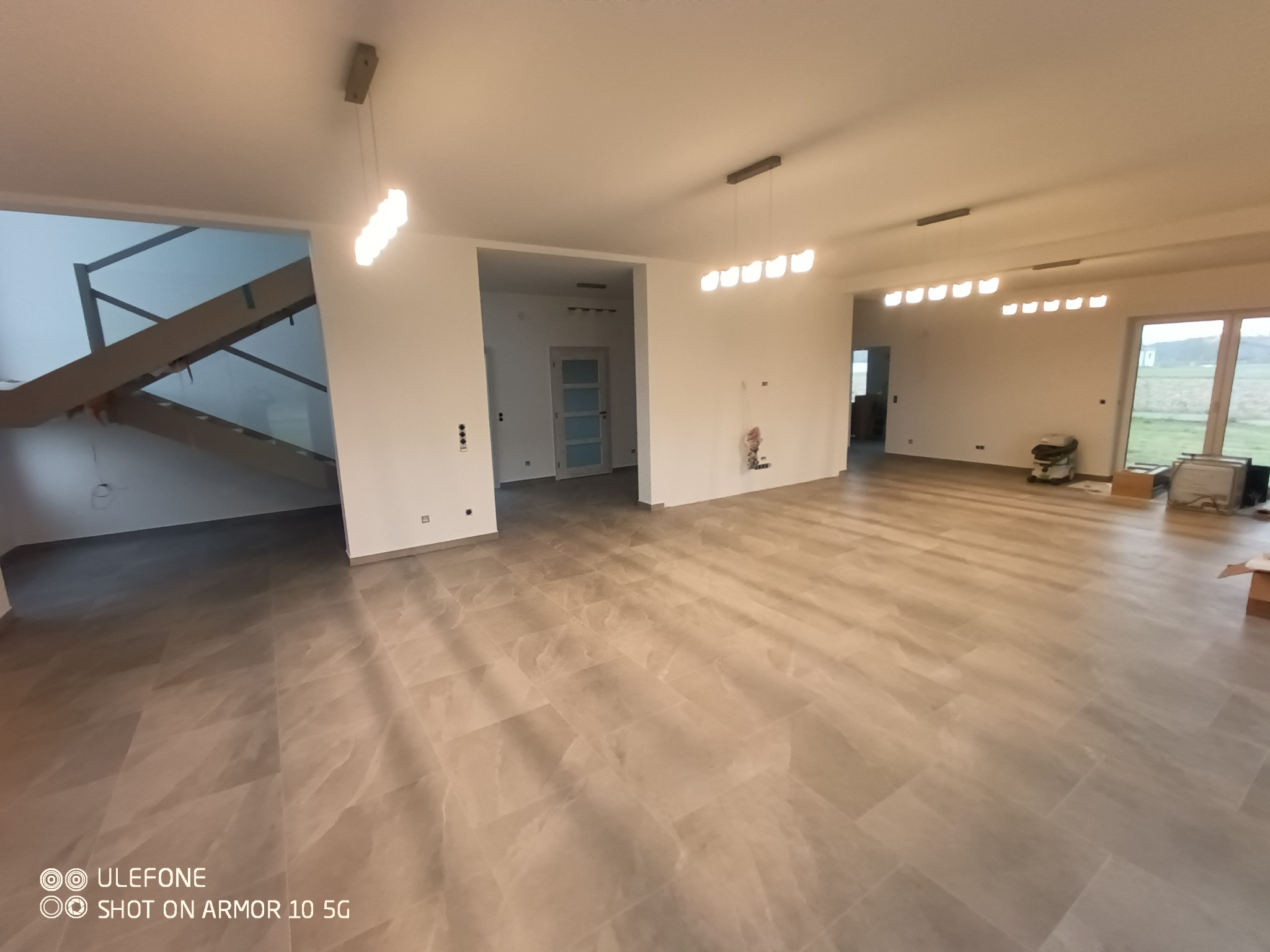 Przestronne wnętrze w stanie deweloperskim z szarą, imitującą kamień podłogą, białymi ścianami, wiszącymi lampami i widokiem na zewnątrz przez duże okno; widoczne przygotowania do dalszych prac...