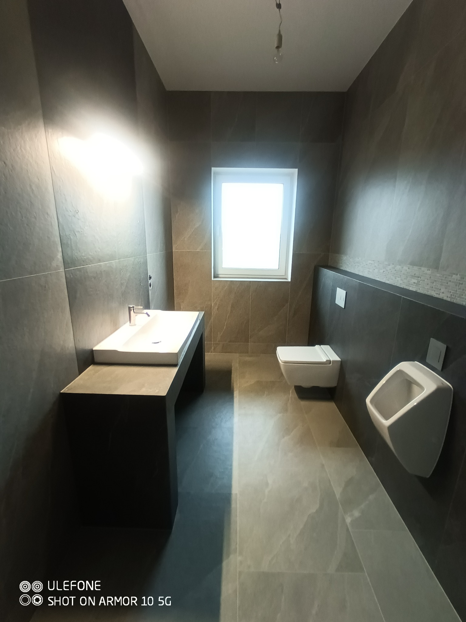 Nowoczesna łazienka z szarymi płytkami imitującymi marmur, wiszącą toaletą, umywalką na czarnym blacie i pisuarze, oświetlona światłem dziennym z okna.