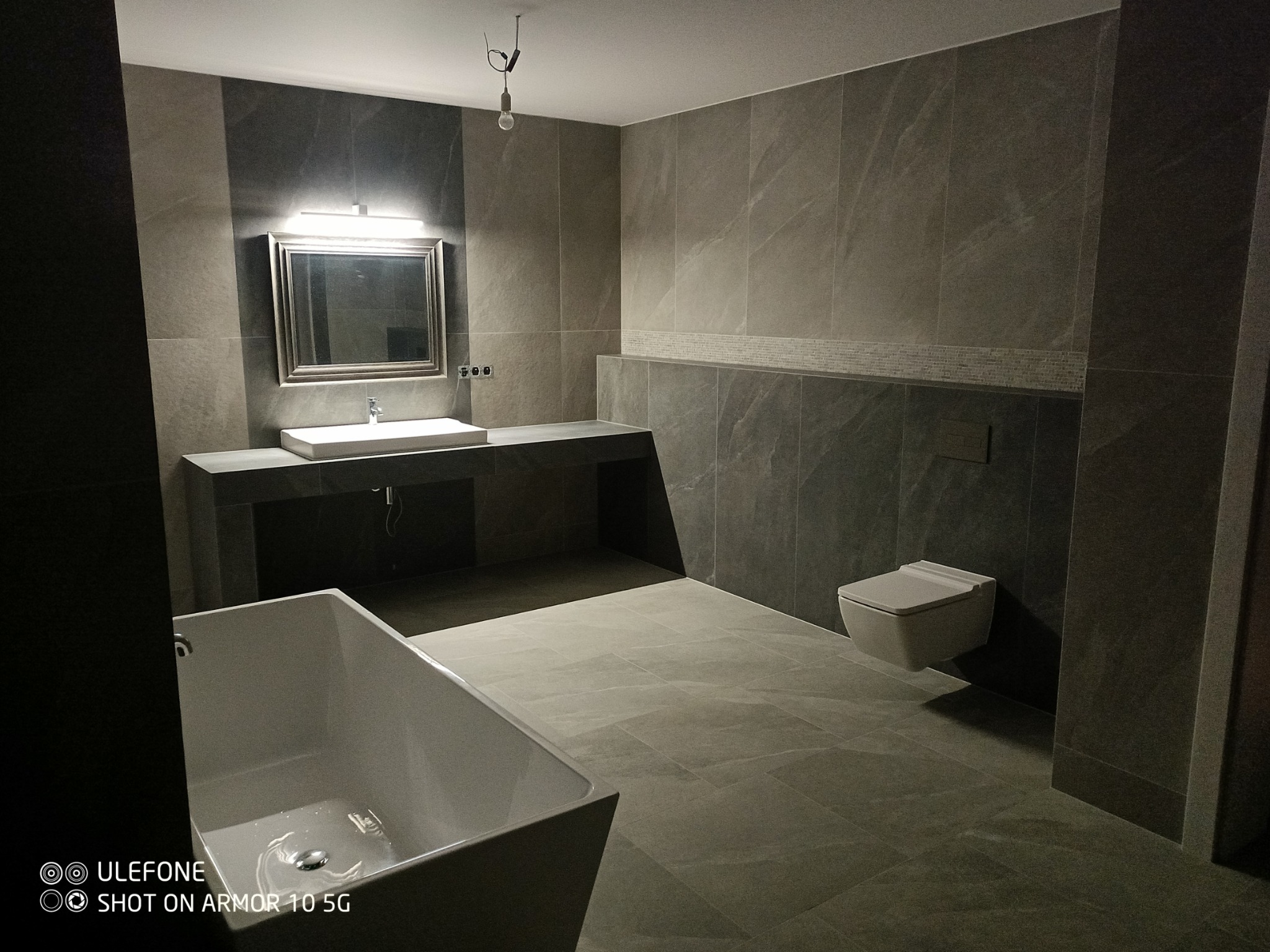 Nowoczesna łazienka w odcieniach szarości z wanną wolnostojącą, podwieszaną toaletą i dużym lustrem z oświetleniem LED nad umywalką.