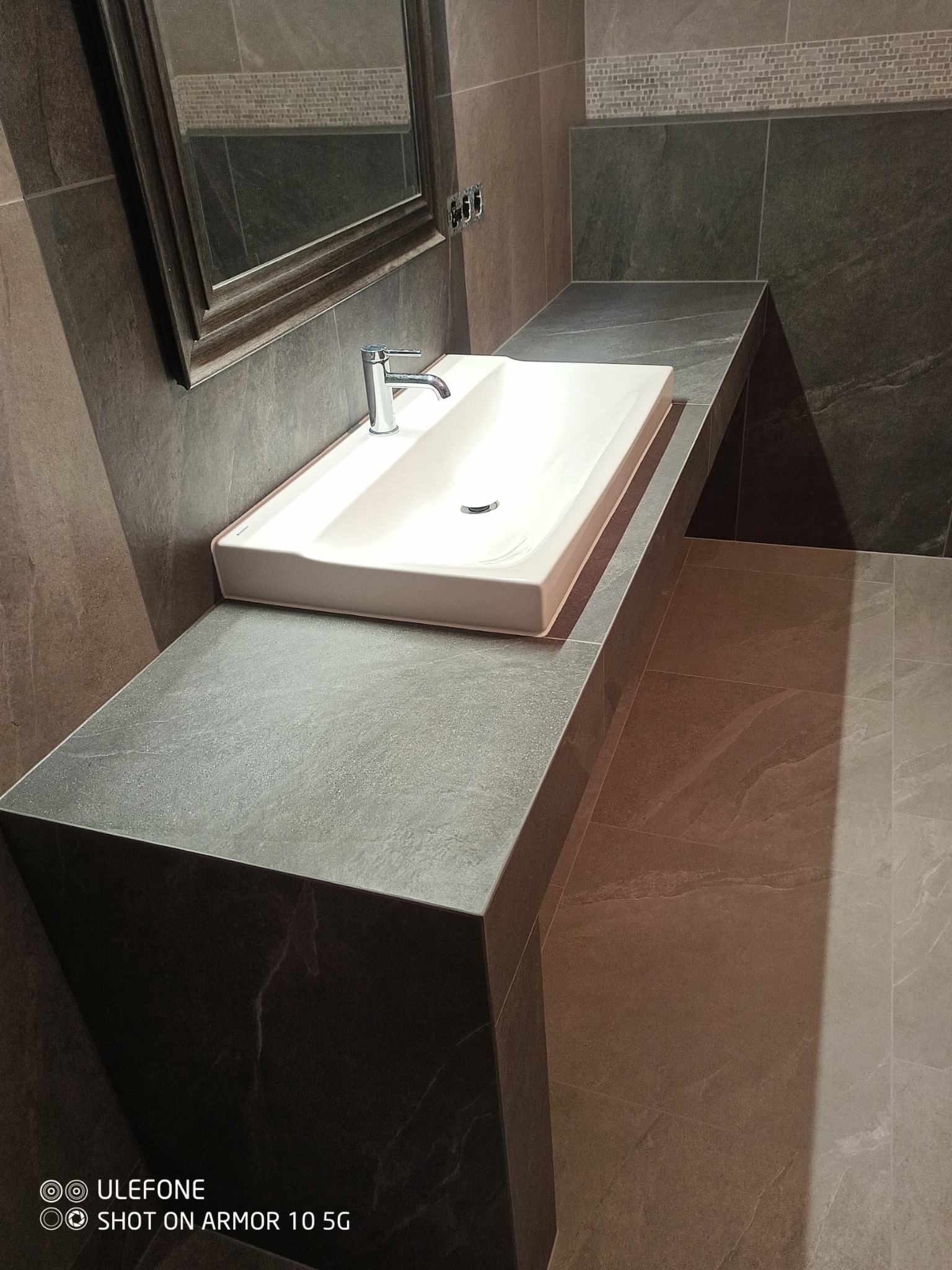 Nowoczesna łazienka z umywalką nablatową, szarym blatem i płytkami imitującymi kamień, lustro z drewnianą ramą.