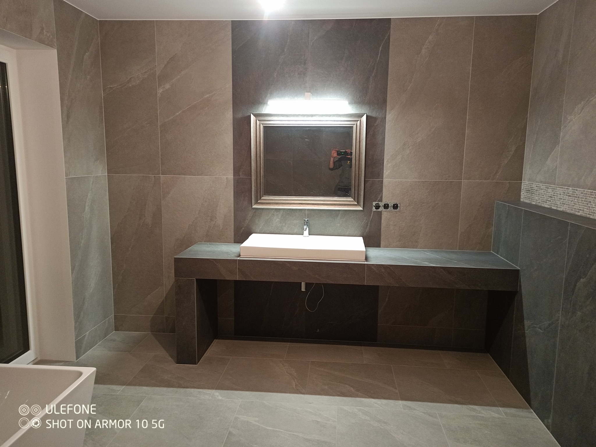 Nowoczesna łazienka z umywalką nablatową, szarymi płytkami imitującymi kamień na ścianach i podłodze, oraz dużym lustrem z oświetleniem LED. Widoczne trzy gniazdka elektryczne pod lustrem.