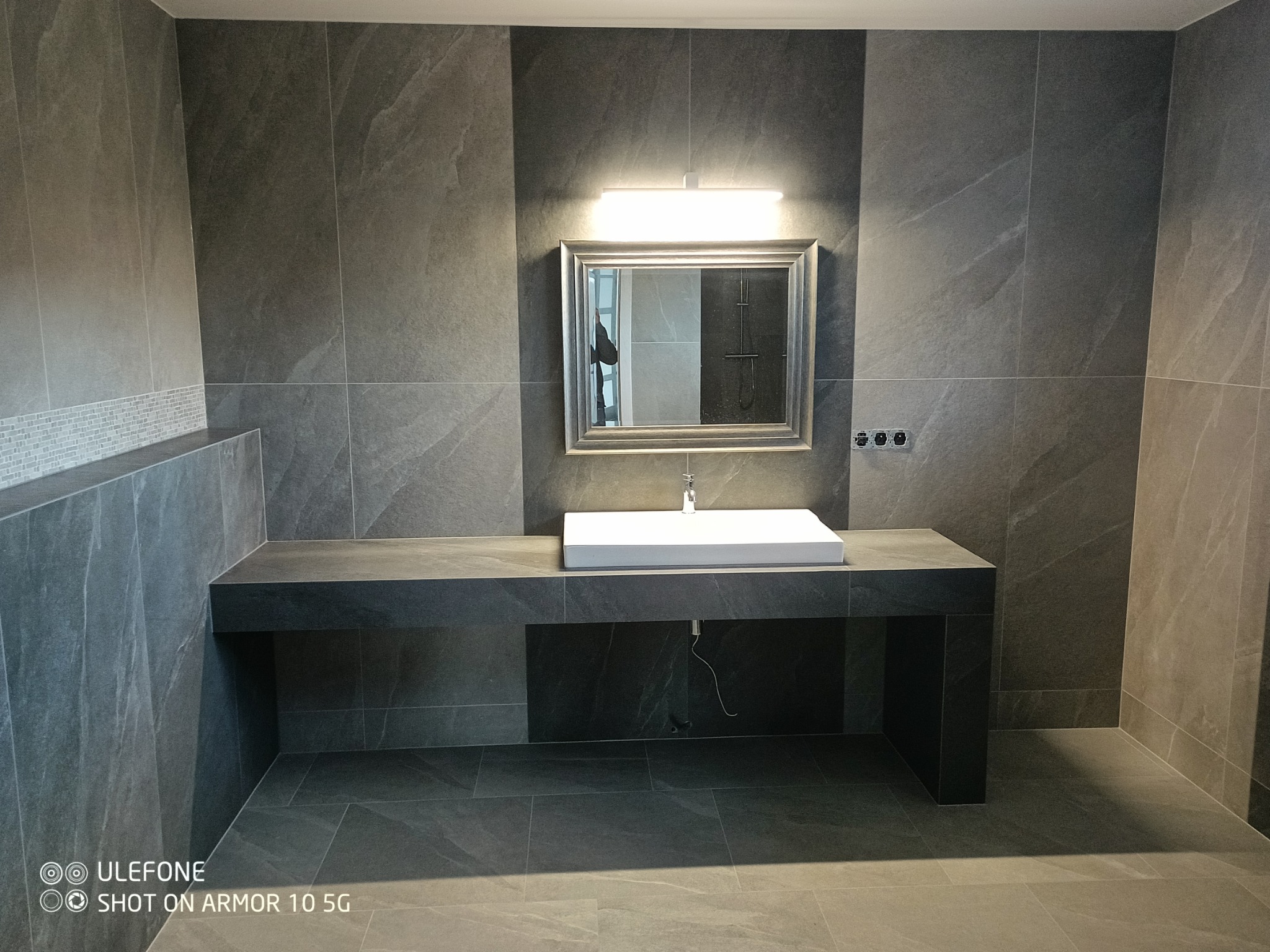 Nowoczesna łazienka w odcieniach szarości z prostokątną umywalką nablatową, lustrem z podświetleniem LED i płytkami imitującymi kamień na ścianach i podłodze.