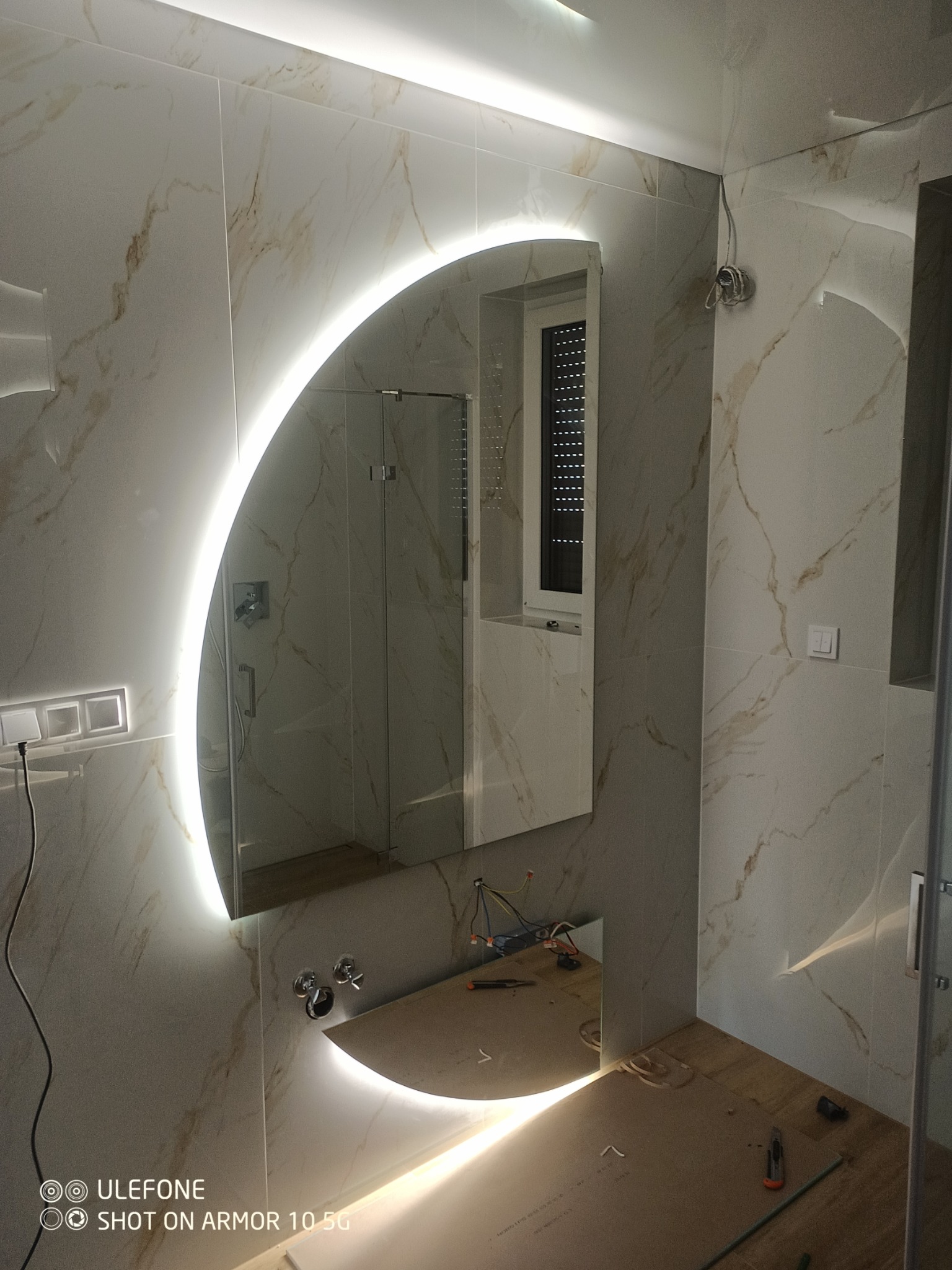 Łazienka w trakcie remontu: marmurowe płytki, podświetlane lustro w kształcie półksiężyca, widoczne przewody i narzędzia na blacie, kabina prysznicowa w tle.