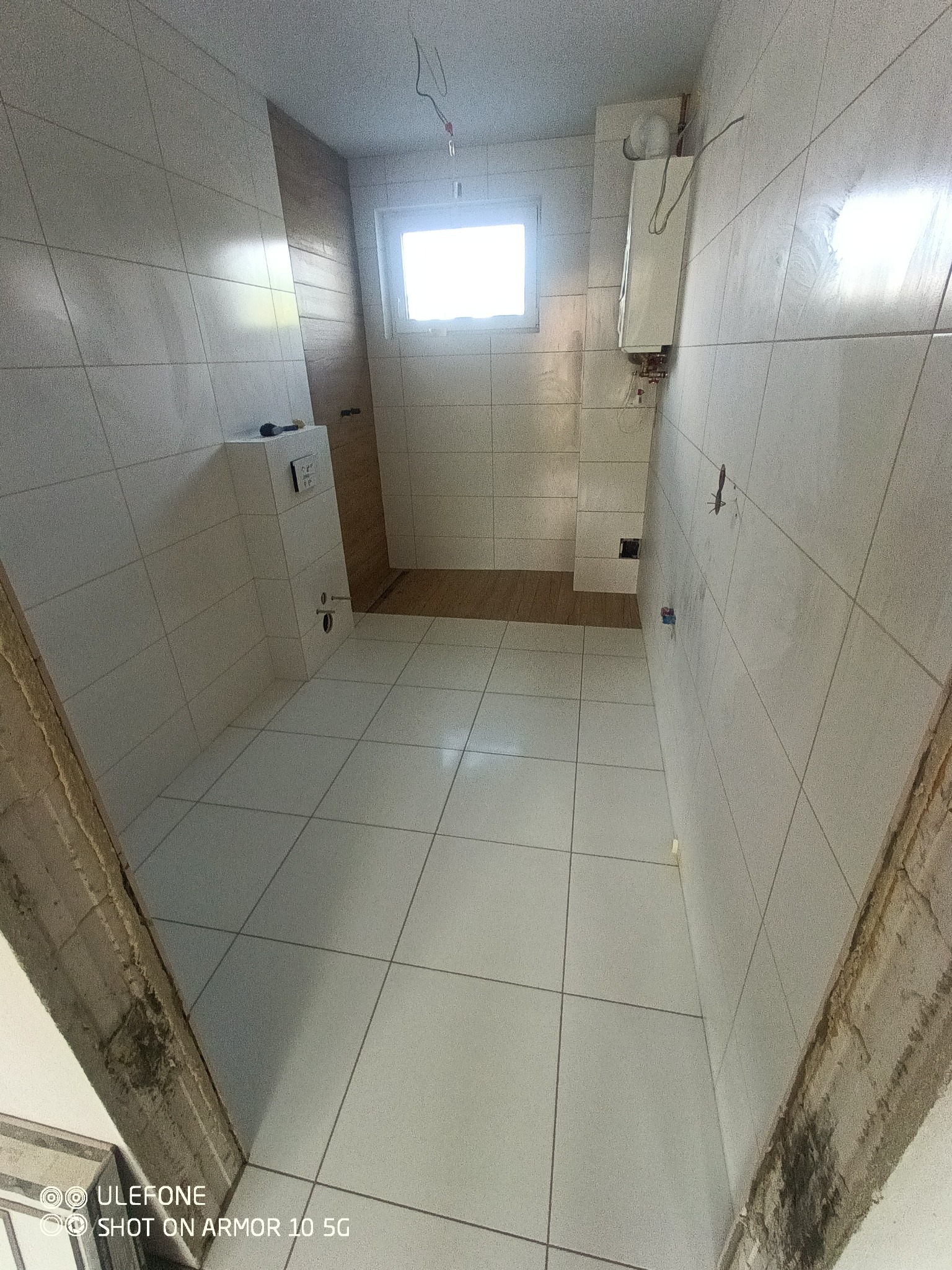 Wnętrze łazienki w trakcie remontu, widoczne białe płytki na ścianach i podłodze, zamontowany stelaż podtynkowy WC, piec gazowy na ścianie, surowe wykończenie ościeżnicy drzwiowej, okno w tle.