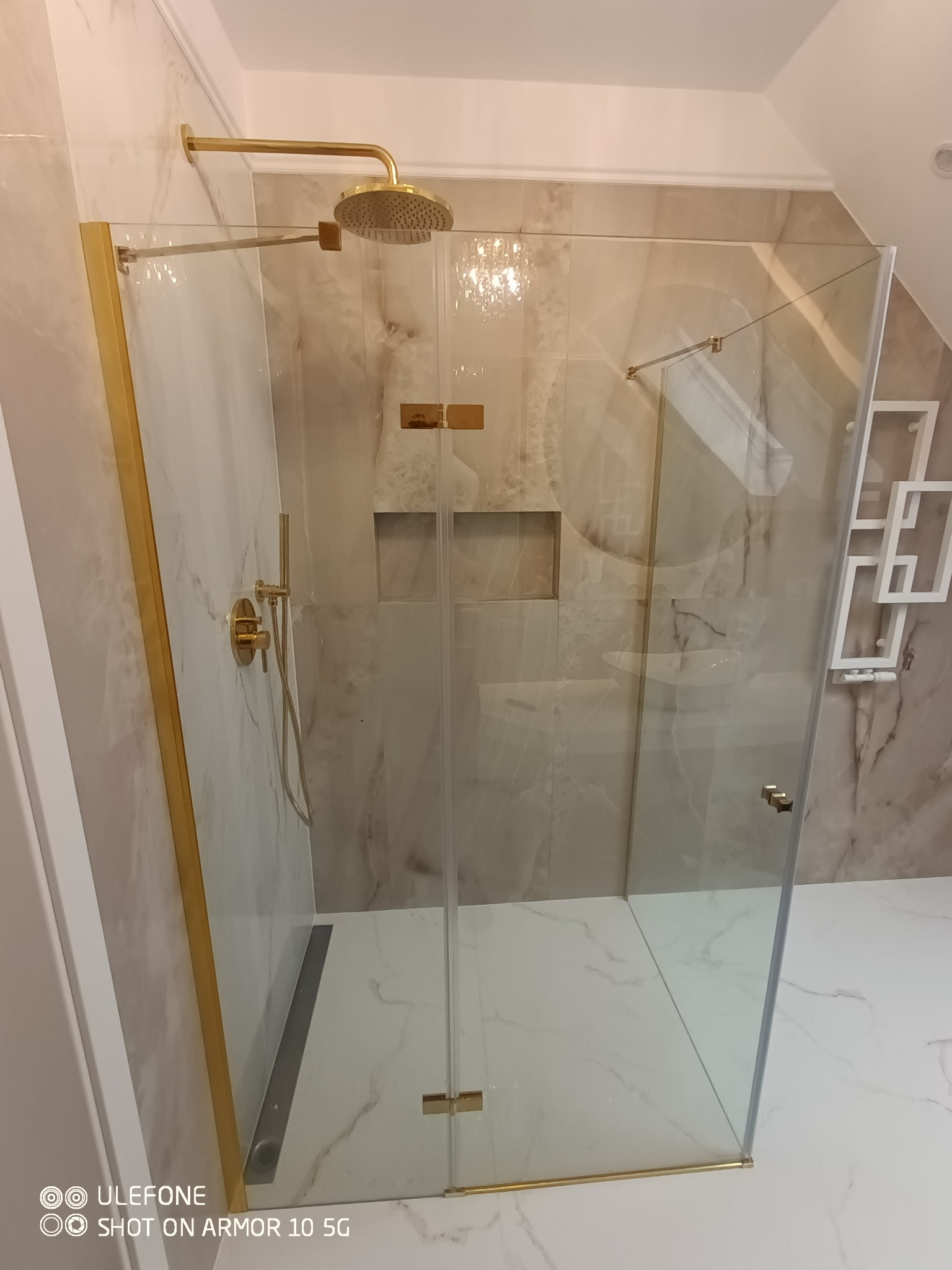 Szklana kabina prysznicowa z złotymi elementami wykończeniowymi, zamontowana w łazience wyłożonej marmurowymi płytkami z wnęką, widoczna deszczownica.