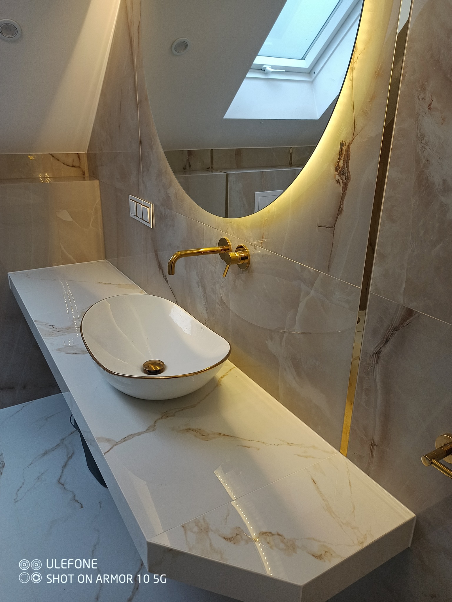 Elegancka łazienka z owalną umywalką nablatową w kolorze białym ze złotymi akcentami, bateria umywalkowa w kolorze złotym, lustro z podświetleniem LED i marmurowe płytki na ścianach