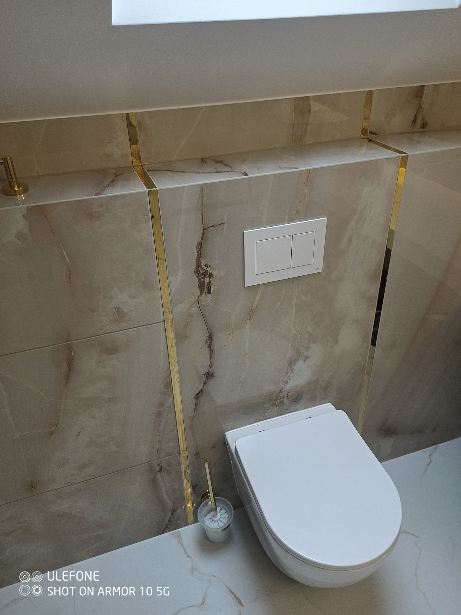 Nowoczesna toaleta podwieszana z beżowymi płytkami imitującymi marmur, wykończona złotymi listwami, widoczny szczotka do WC w złotym uchwycie