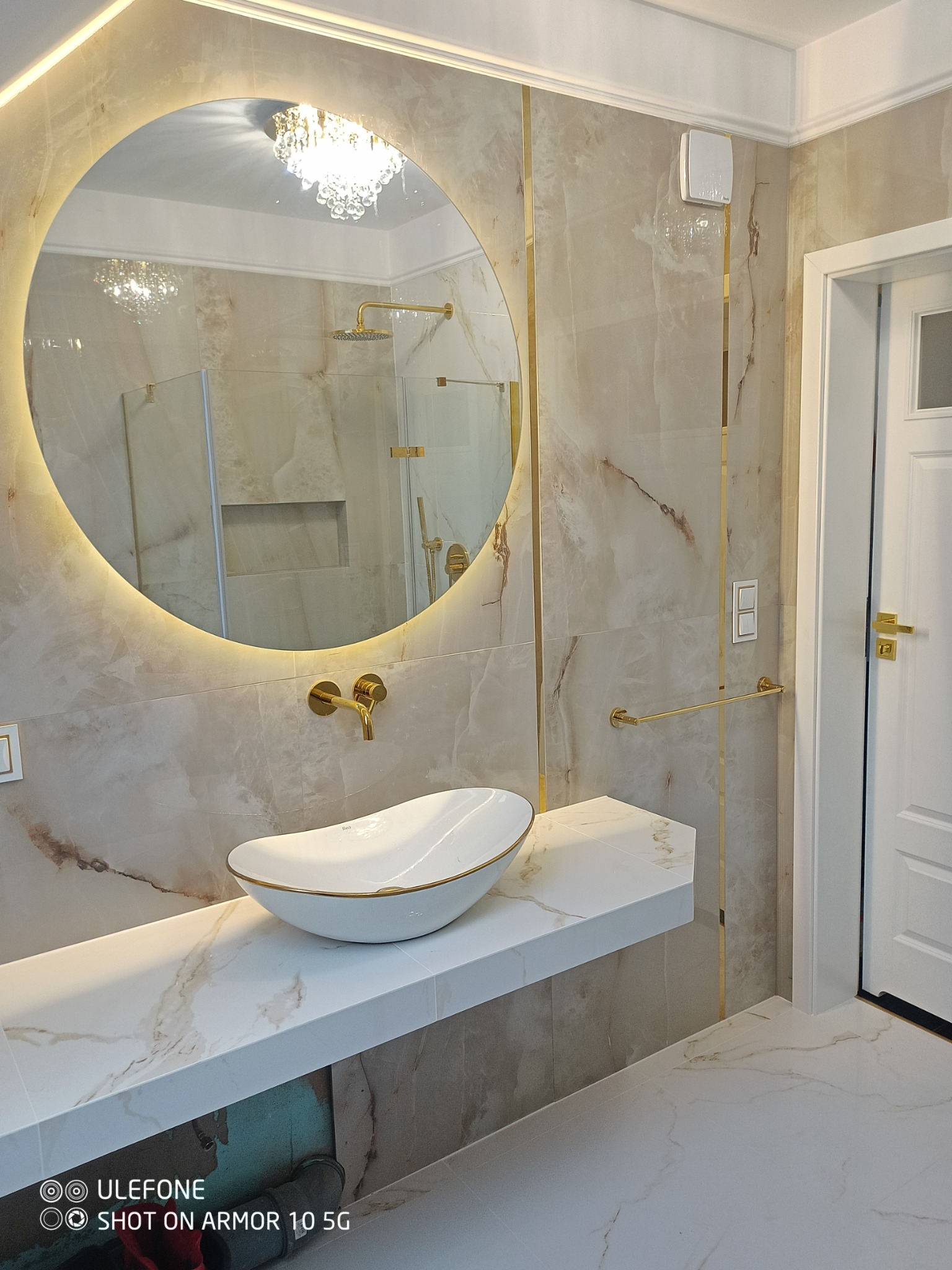Elegancka łazienka z owalną umywalką nablatową z złotym rantem, podświetlanym okrągłym lustrem i marmurowymi płytkami z złotymi akcentami.
