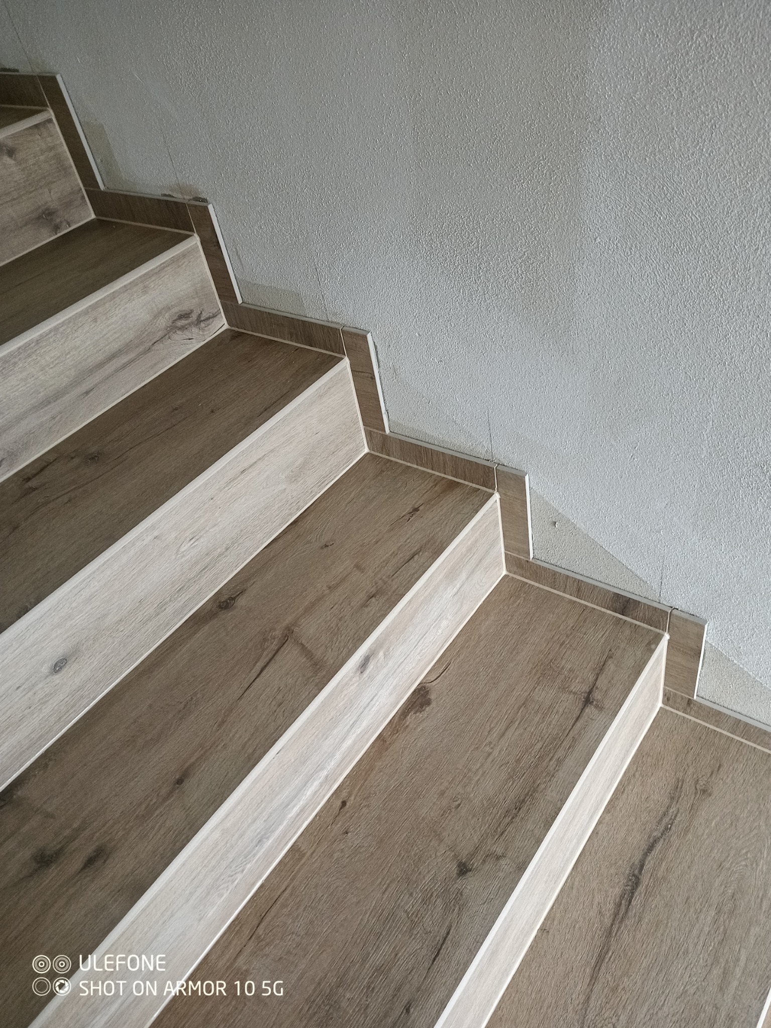 Schody wykończone panelami imitującymi drewno, z listwami przypodłogowymi i fragmentem ściany o chropowatej fakturze.