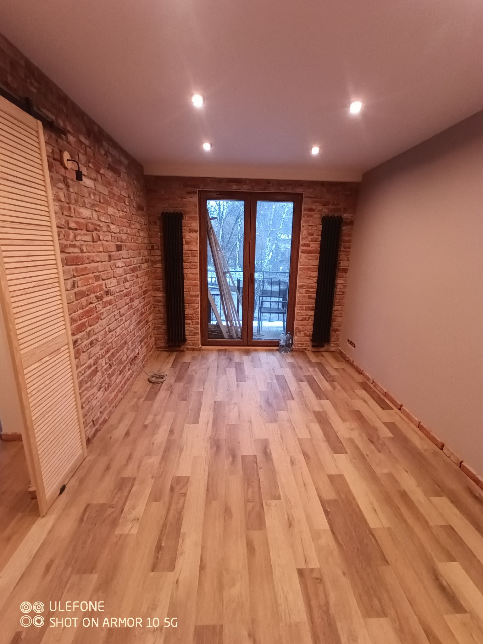 Wykończone wnętrze pokoju z panelami podłogowymi, ścianą z cegły, grzejnikami żeberkowymi i drewnianymi drzwiami balkonowymi z widokiem na zewnątrz.