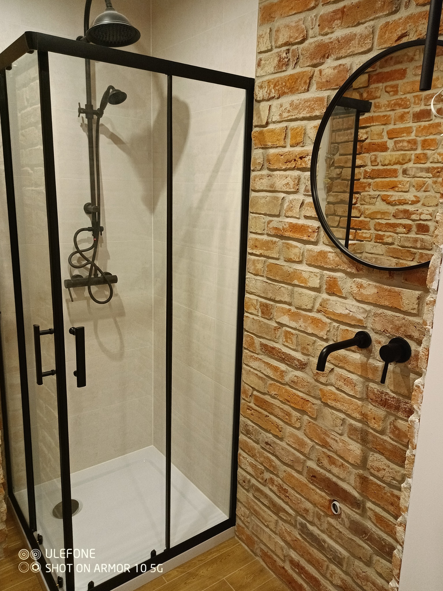 Kabina prysznicowa z czarnymi ramami i armaturą na tle ściany z cegły, okrągłe lustro w czarnej ramie, drewnopodobna podłoga.