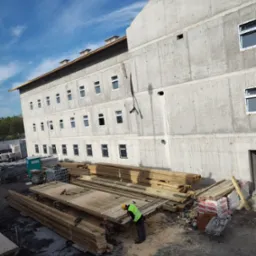 Budowa budynku z betonu architektonicznego: pracownik w kamizelce odblaskowej obrabia drewniane elementy szalunku na tle surowej betonowej konstrukcji wielopiętrowego budynku z oknami.