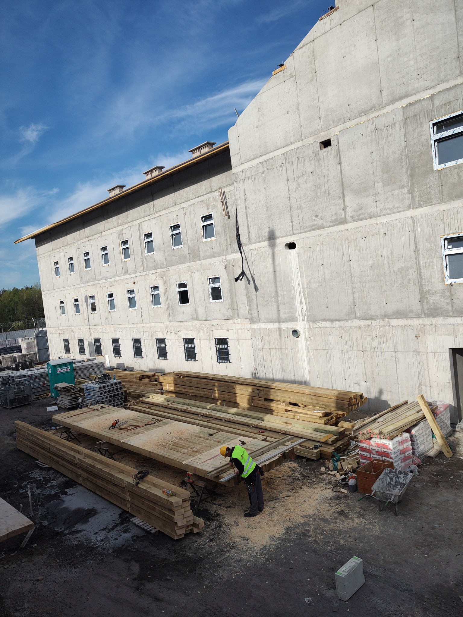 Budowa budynku z betonu architektonicznego: pracownik w kamizelce odblaskowej obrabia drewniane elementy szalunku na tle surowej betonowej konstrukcji wielopiętrowego budynku z oknami.